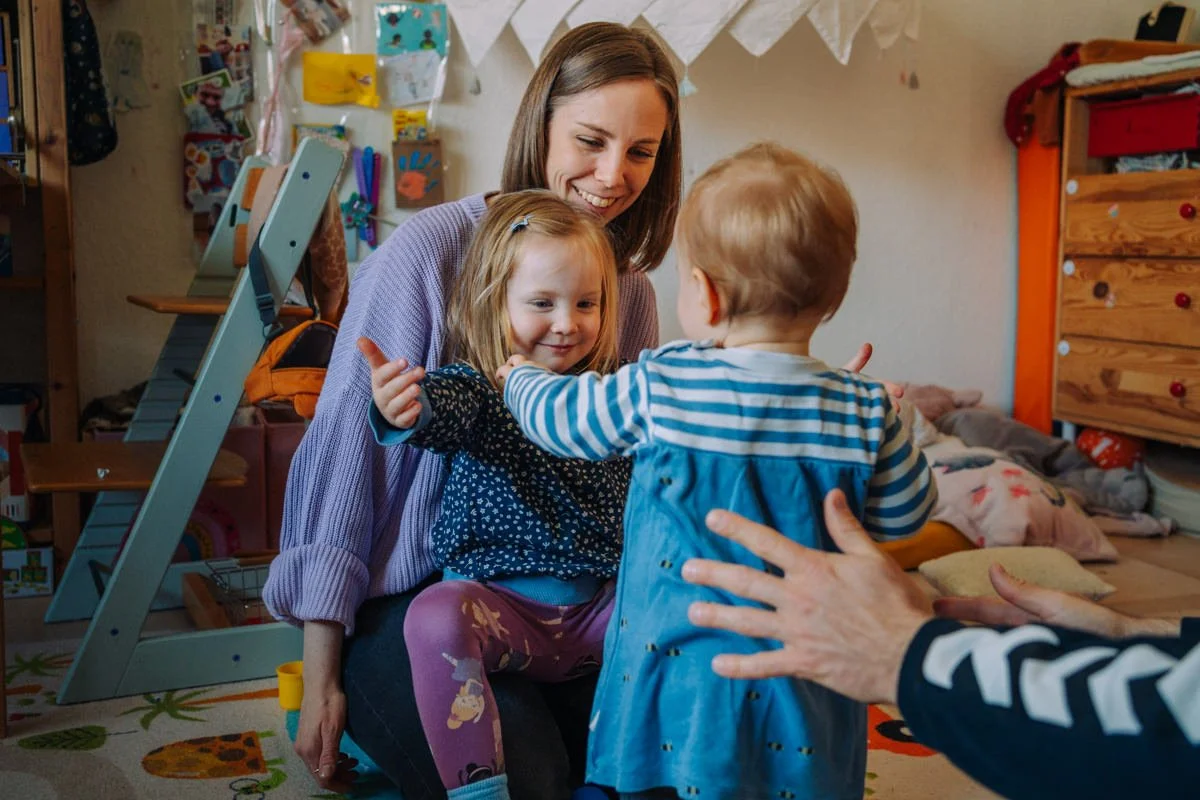 Eine Frau mit zwei Kindern in einem Kinderzimmer, die sich umarmen und lachen, während gespielt wird.