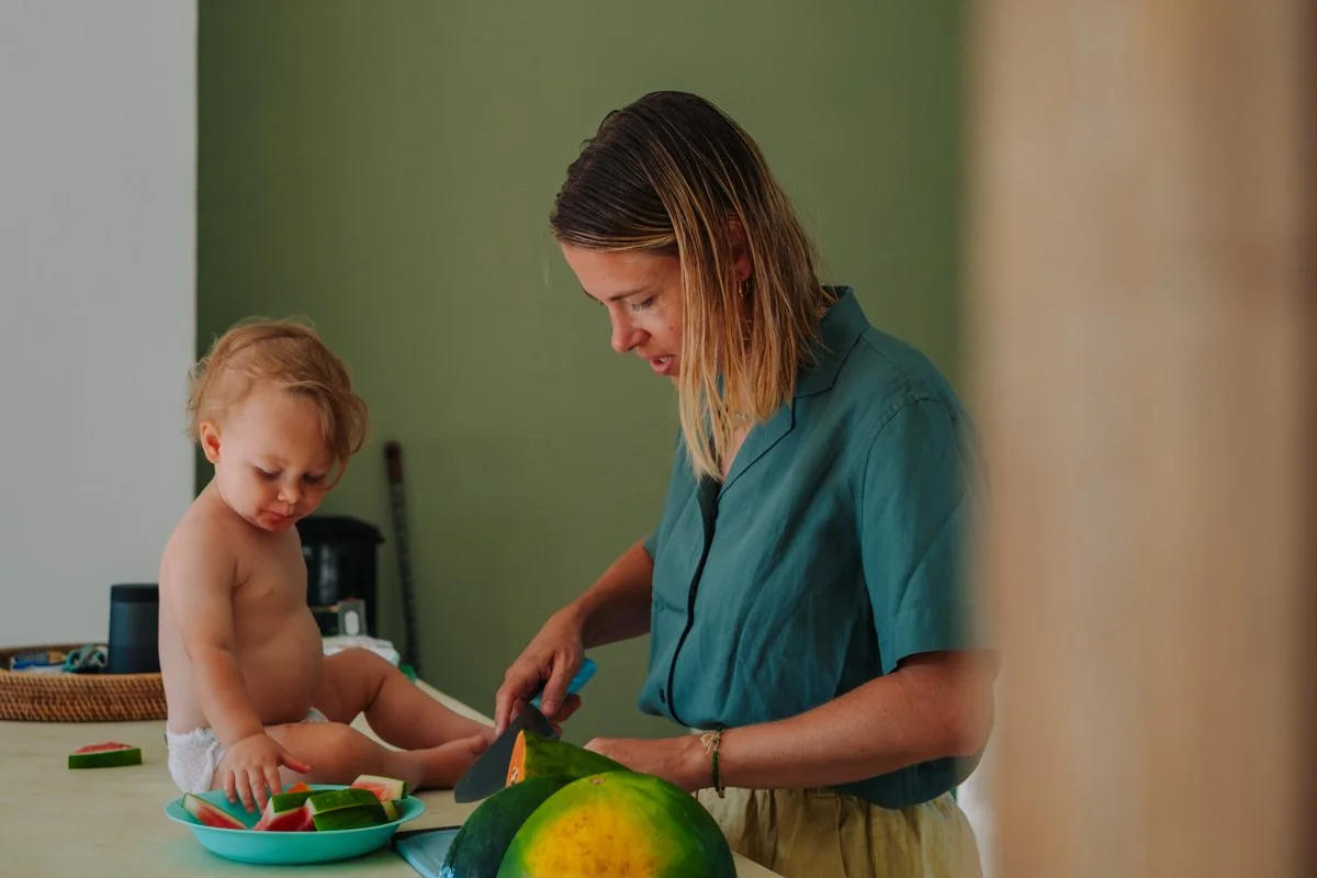Eine Frau schneidet Wassermelone in einer Küche, neben einem kleinen nackten Kind, das auf einem Tisch sitzt.