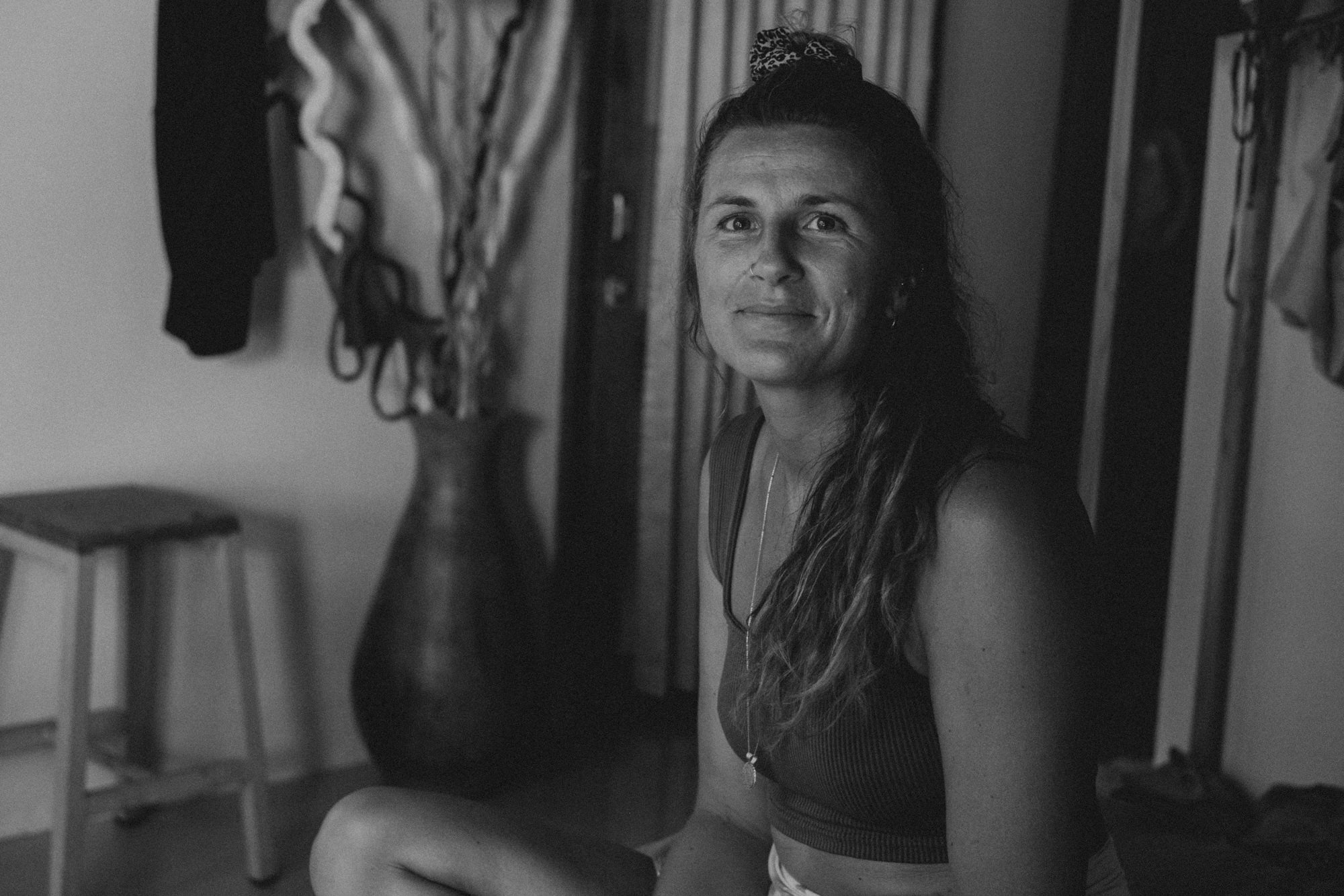 Schwarz-weiß-Foto einer lächelnden Frau mit langen, lockigen Haaren, die auf einem Stuhl sitzt, im Hintergrund eine Vase mit großen Zweigen und eine Kommode.