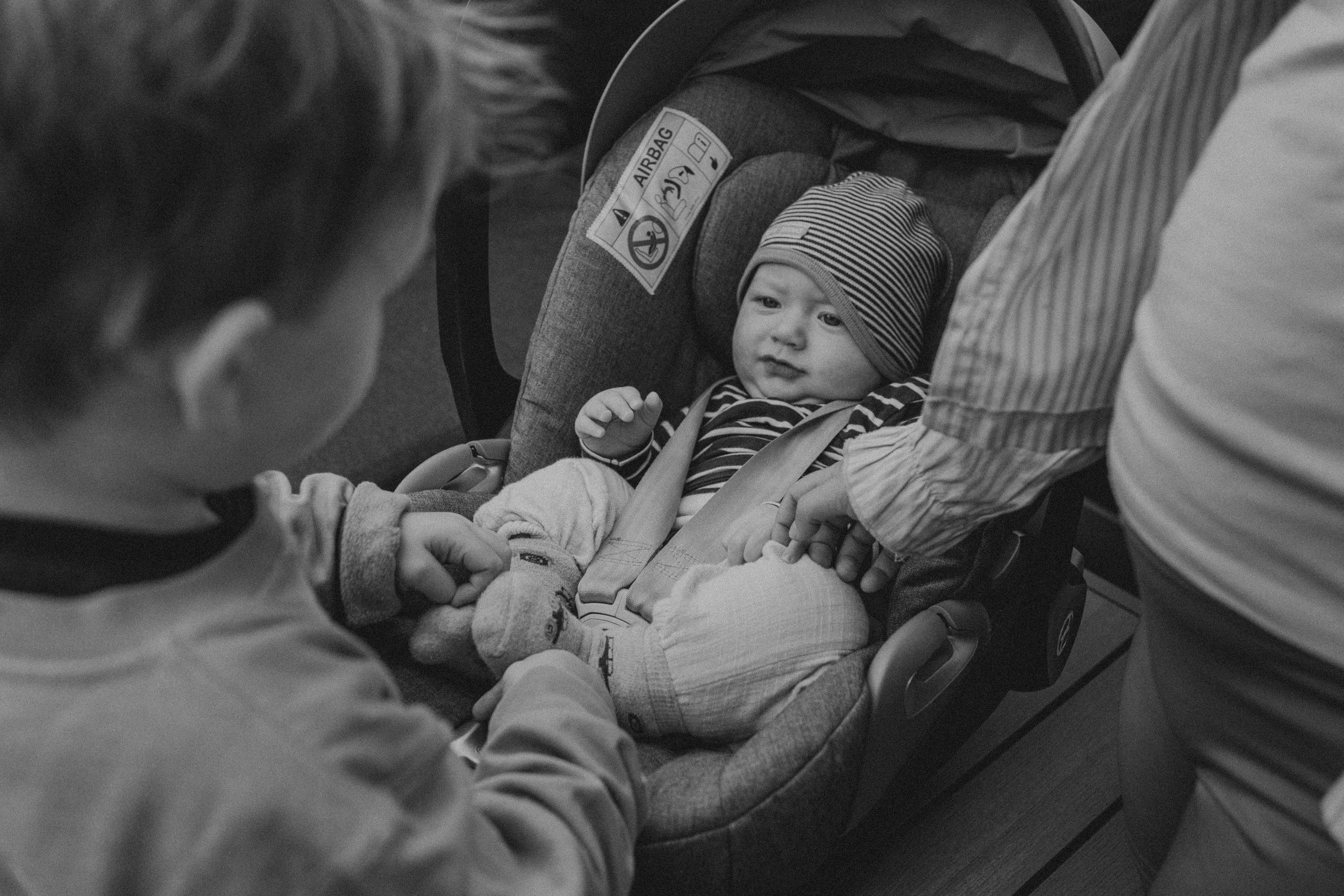 Ein Baby sitzt in einem Autositz, umgeben von Erwachsenen, und schaut neugierig.