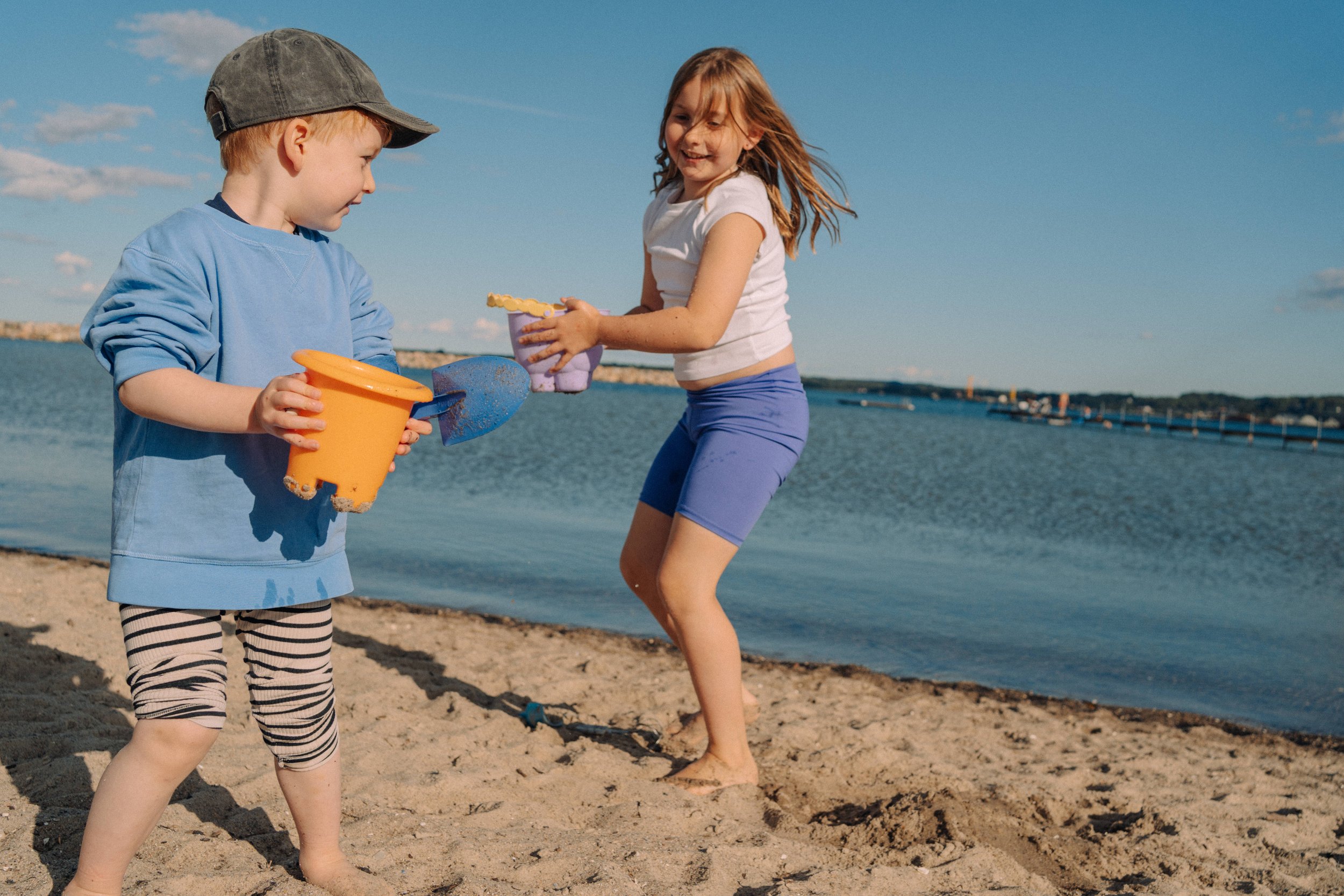 Zwei Kinder spielen im Sand am Wasser, ein Junge mit blauer Jacke und Mütze, ein Mädchen in weißem T-Shirt und blauen Shorts, mit Spielzeug am Strand.