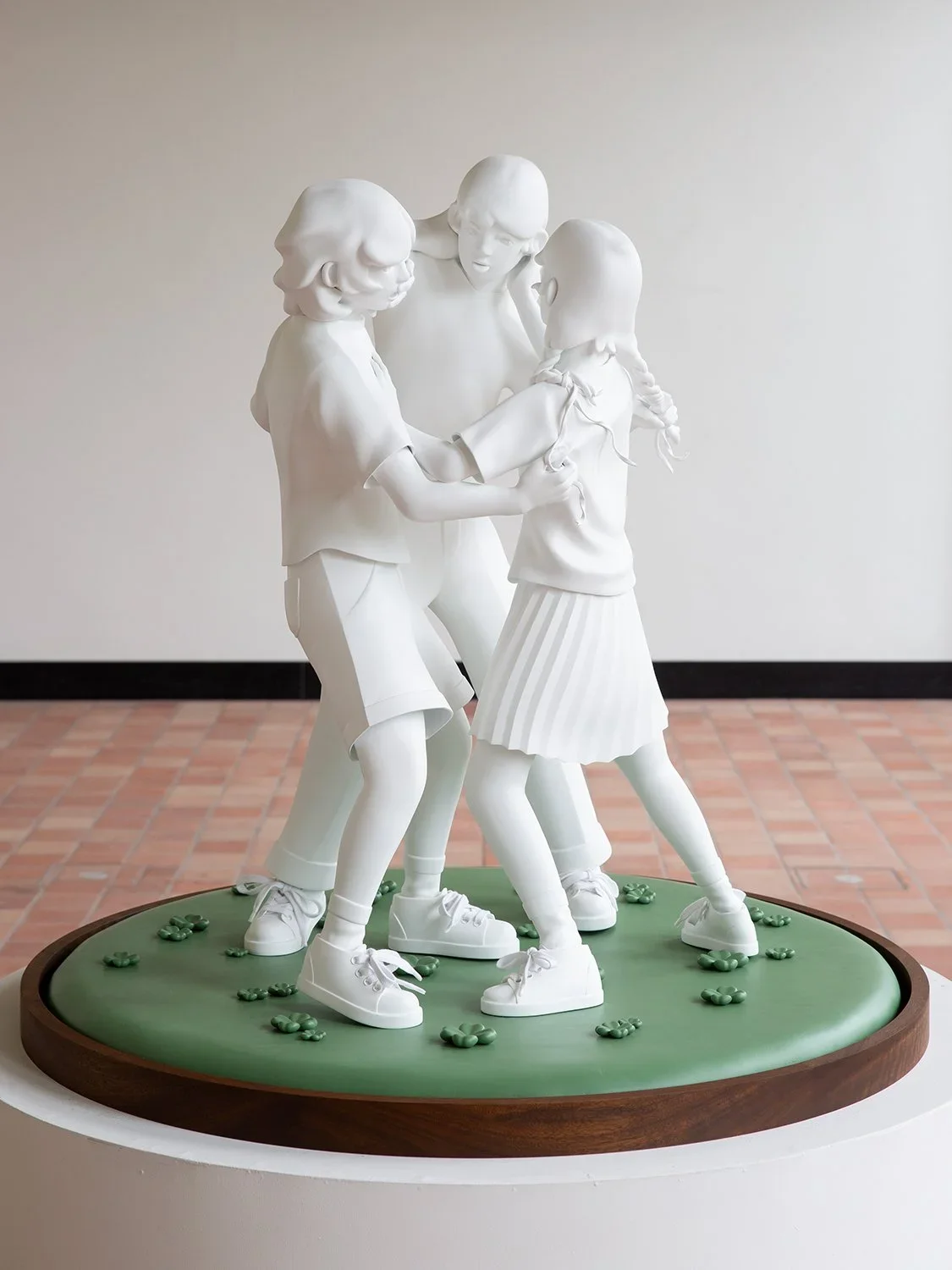 A white sculpture of four children holding hands and playing together on a round green base with small green flowers, displayed indoors against a plain wall.