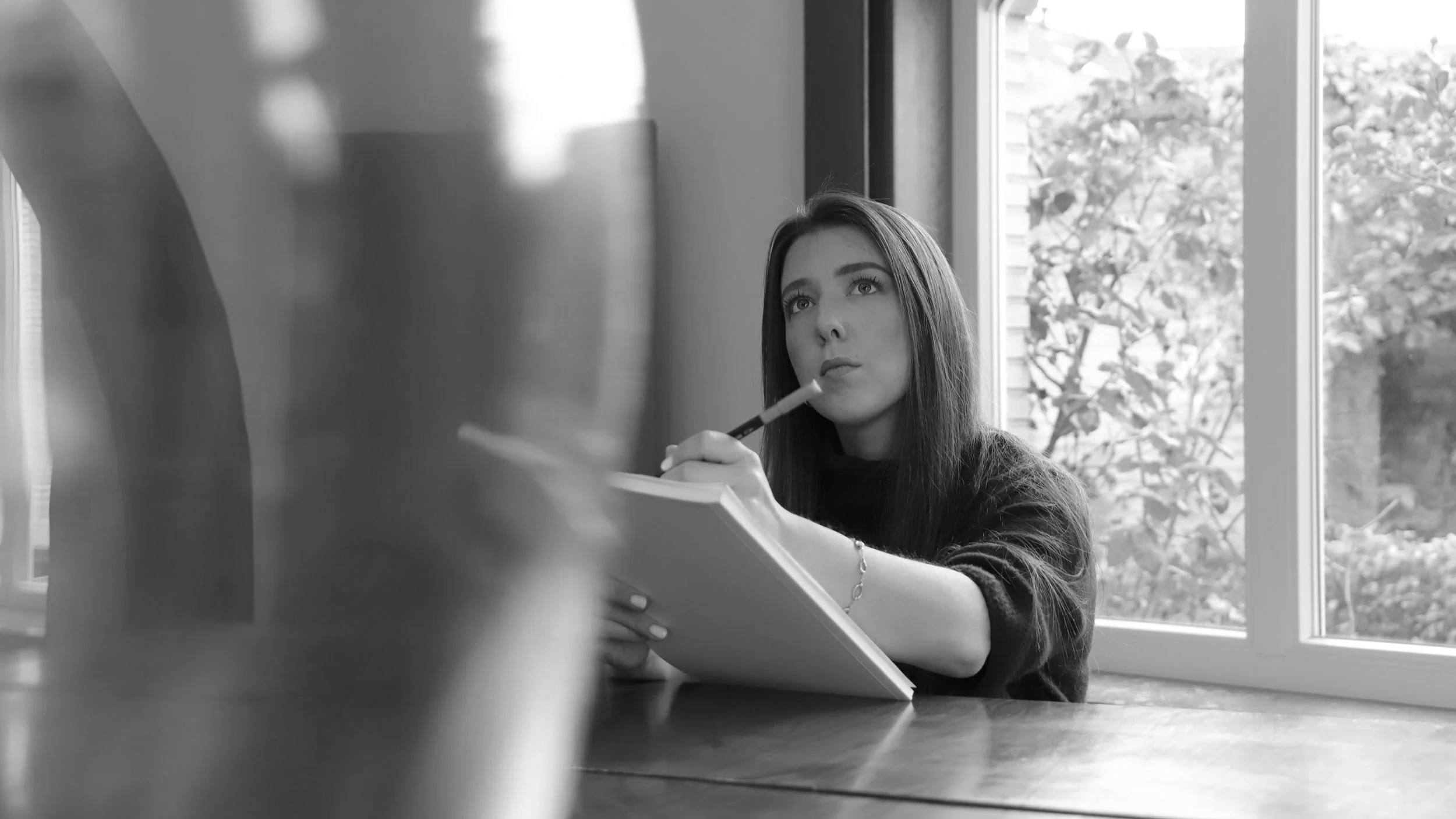 A young woman with long hair sits at a wooden table, holding a notepad and a pen, with a thoughtful expression as she looks out a window displaying trees outside.