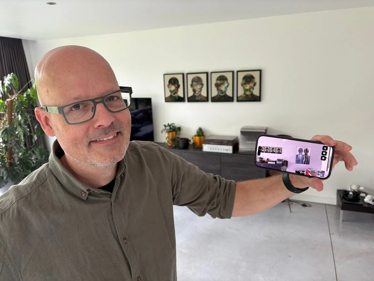 A man with glasses and a beard taking a selfie in a modern living room, holding a smartphone with the camera facing him. The background features a TV, framed artwork, potted plants, and a console.