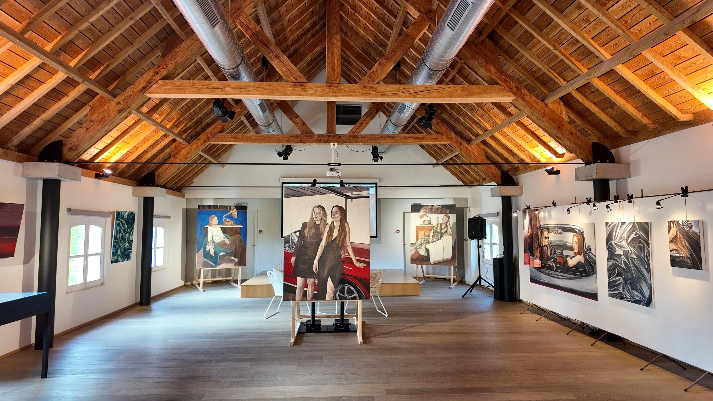 Art gallery with paintings of people and objects displayed on white walls and on easels, with a wooden ceiling and large windows.