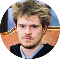 A young man with messy, blonde hair and a serious expression, sitting on a wooden chair with a blue background.