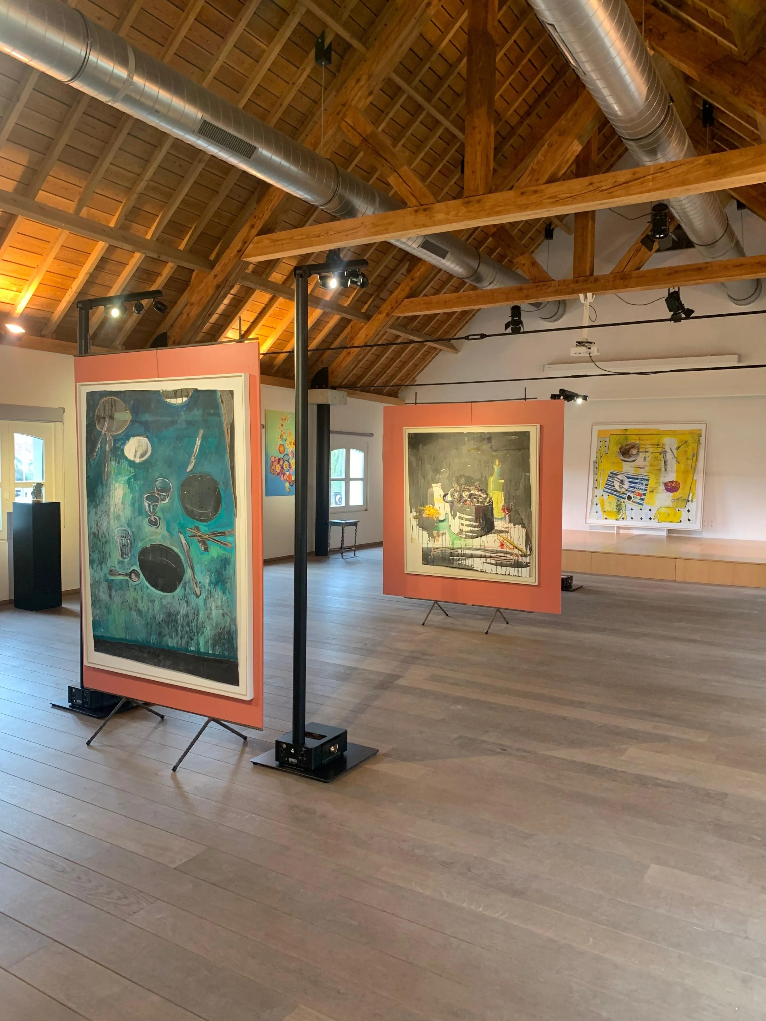 Interior of an art gallery with wooden ceiling beams, ductwork, and artwork displayed on free-standing boards.