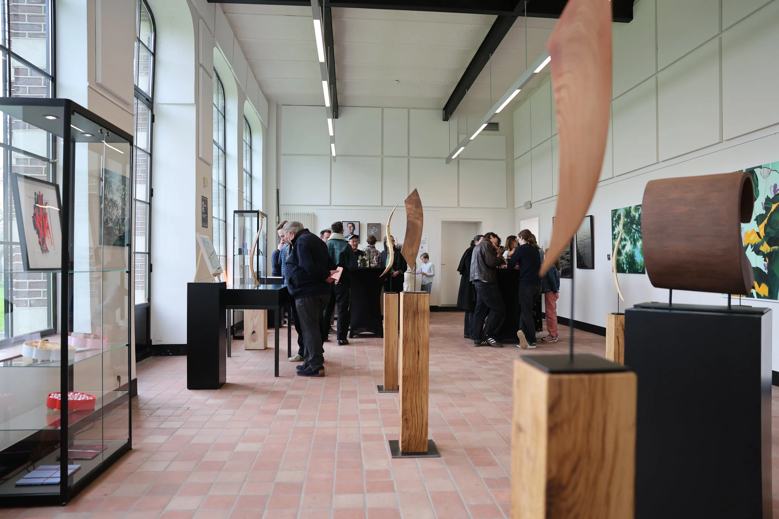 Mensen die kunst bekijken in een gallery met grote ramen, schilderijen aan de muren en houten sculpturen.