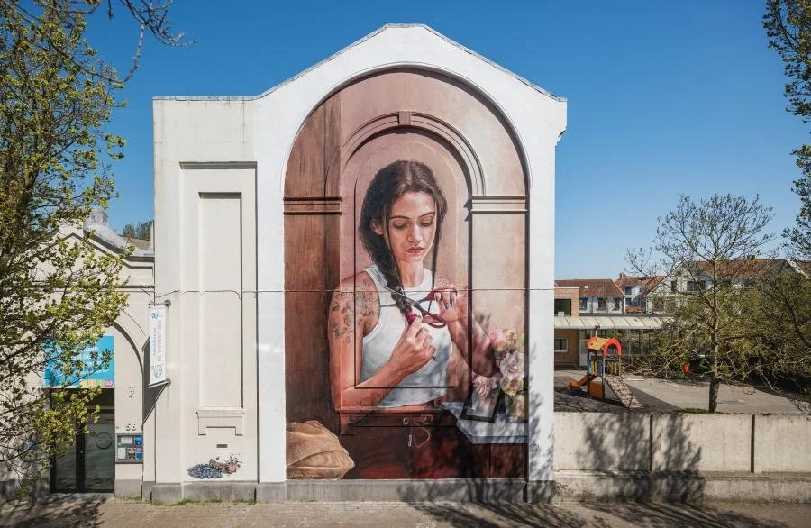 A large mural of a woman with dark hair, wearing a white tank top, looking at a pair of glasses she is holding in her hands. She has tattoos on her arms and is surrounded by a wooden frame illustration within the mural. The mural is on the side of a white building, with trees and a playground visible in the background on a clear day.