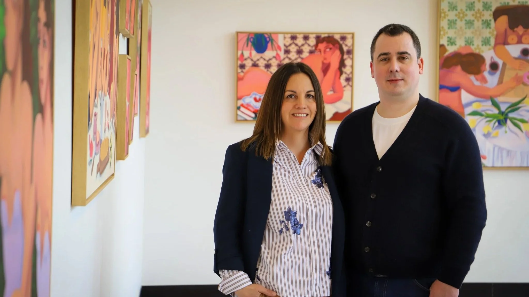 A woman and a man standing side by side in an art gallery, smiling at the camera, with colorful paintings on the wall behind them.