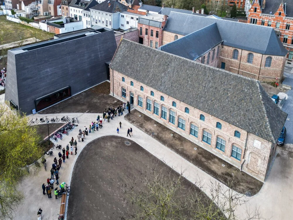Een groep mensen staat in een rij voor een nieuw gebouw of school met een voormalige industriële structuur, omgeven door een kleine tuin en een parkeerplaats met fietsen.