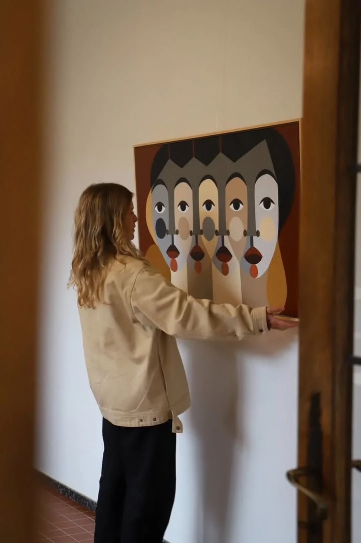 A woman with wavy blonde hair wearing a beige jacket and black pants is hanging a modern abstract painting on a white wall inside a room.