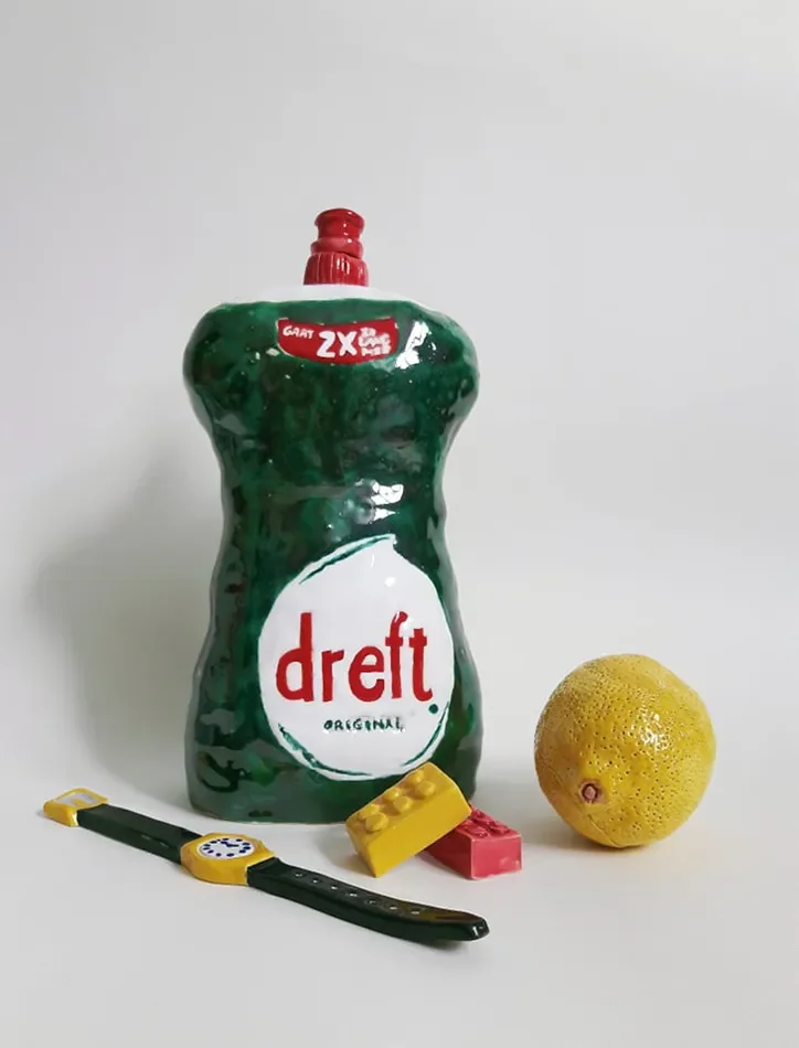 A ceramic bottle of Dreft original liquid detergent, a lemon, a black and yellow wristwatch, and two small LEGO bricks in yellow and red on a white surface.