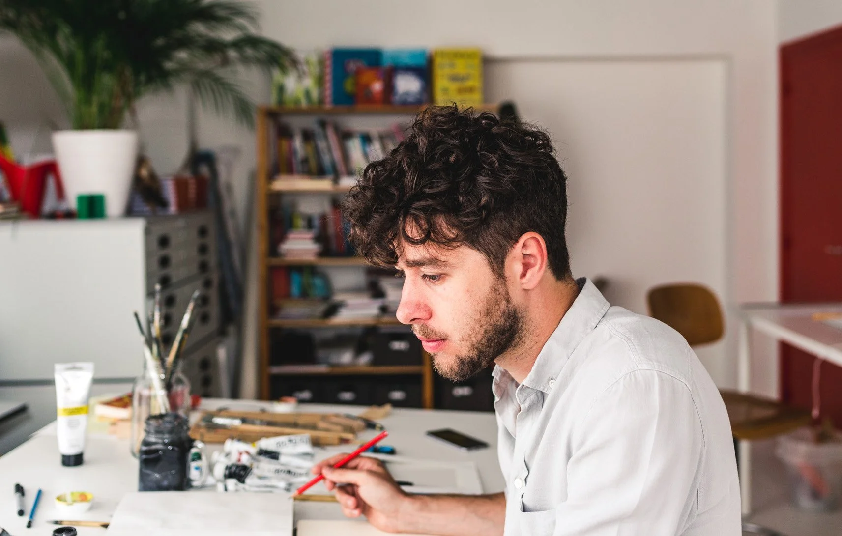 A young man with curly dark hair and a beard sitting at a table in a creative workspace, holding a red pencil, with art supplies and scissors on the table, and a bookshelf filled with books in the background.