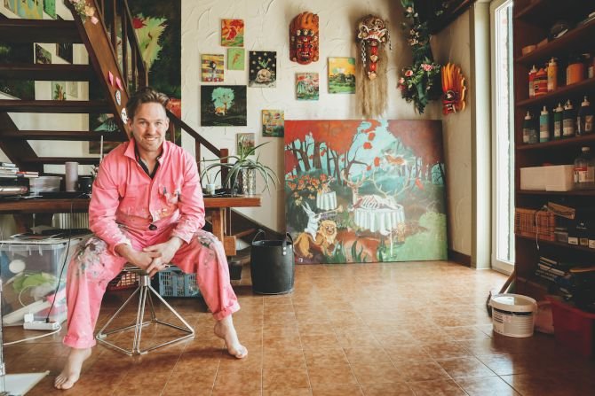 A man sitting on a stool in a room decorated with various colorful artwork, plants, and Asian masks, with a large landscape painting resting against the wall, and shelves filled with art supplies. The room has a window with sunlight coming in and tiled flooring.