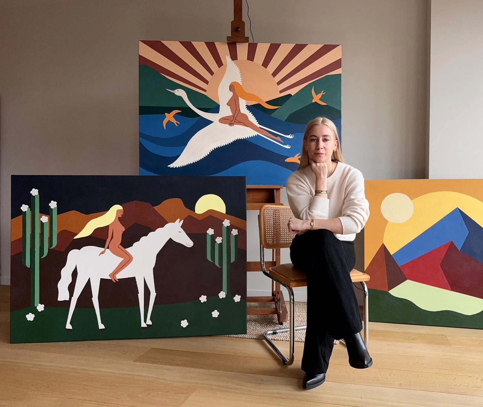 A woman sitting on a chair in front of three colorful landscape paintings, with a large painting of a woman riding a white bird in the background.