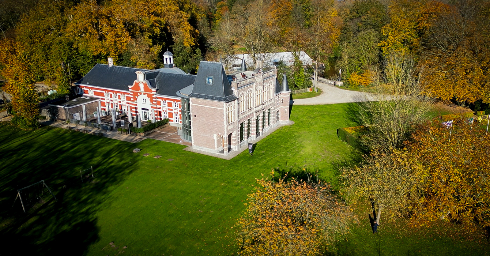 An aerial view of a historic castle-like building surrounded by a lush green lawn and colorful autumn trees.
