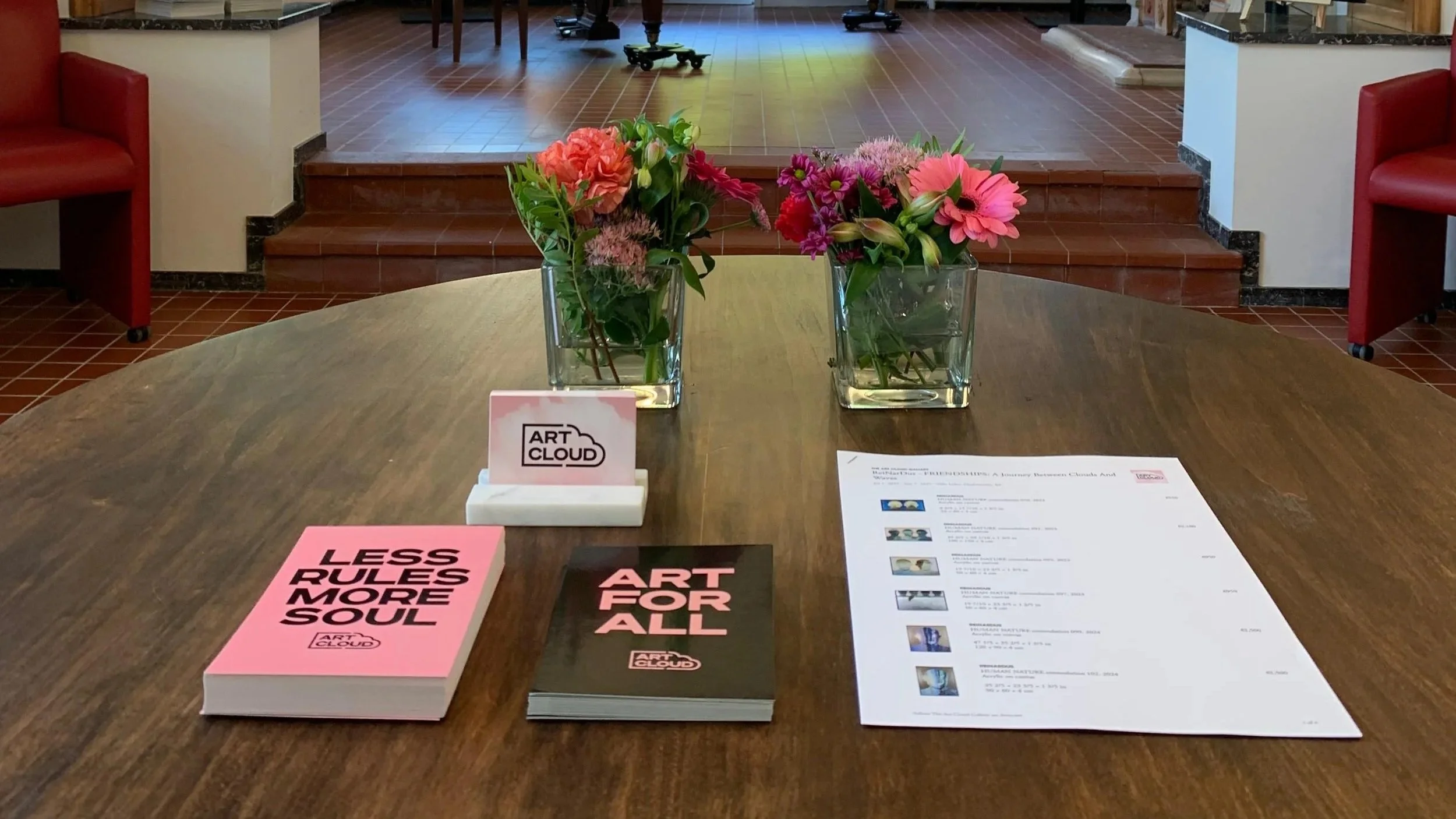 A wooden table with two glass vases of pink and red flowers, a pink booklet titled 'LESS RULES MORE SOUL', a black booklet titled 'ART FOR ALL', a white cardholder with a sign reading 'ART CLOUD', and a white sheet of paper with text and images. In the background, there are red chairs, a tiled floor, and a slight staircase leading to an elevated area.