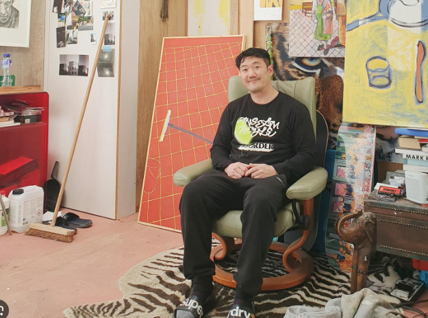 A man sitting in a green armchair in a cluttered room filled with art supplies, paintings, and photos on the wall.
