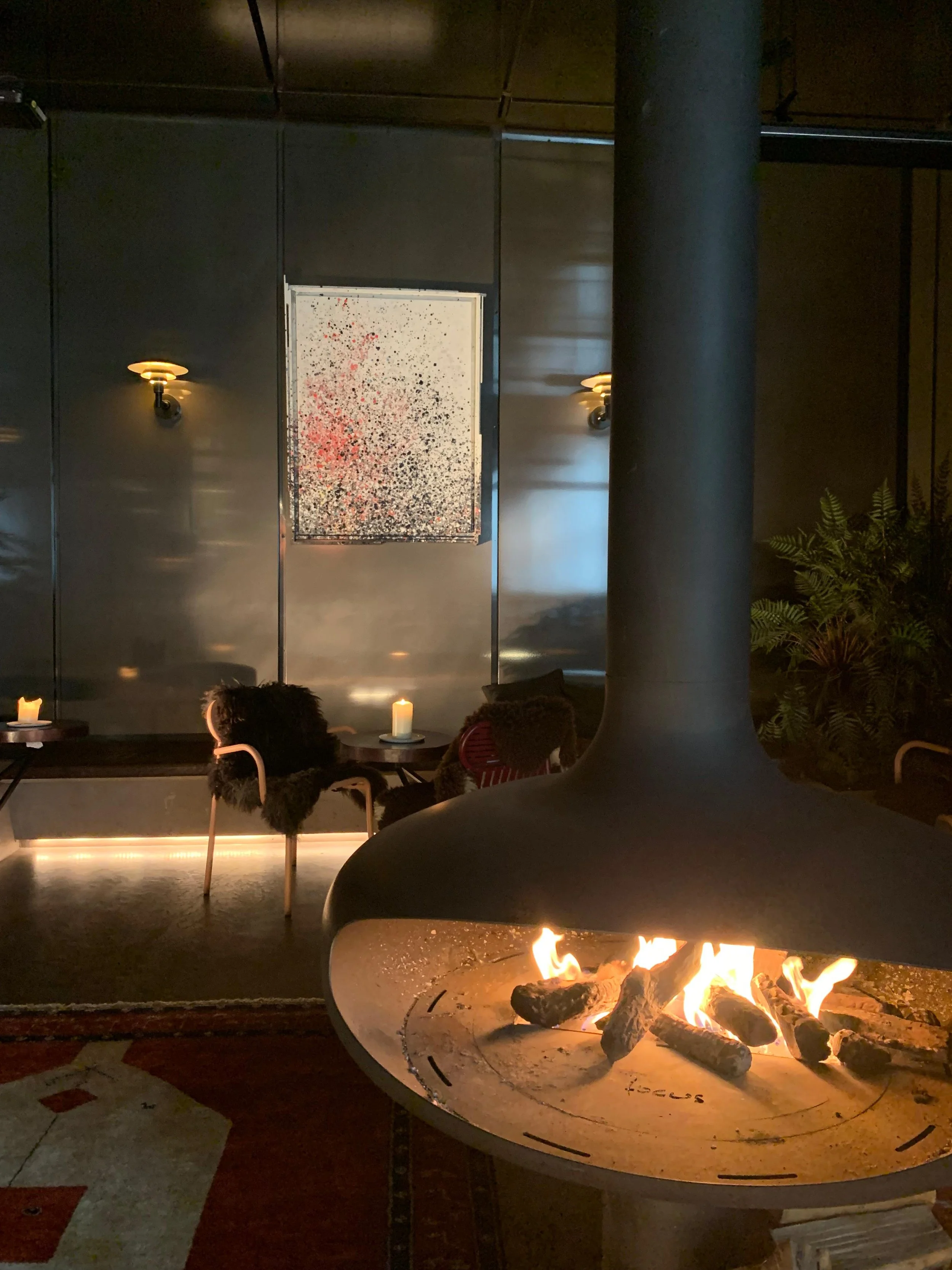 Cozy indoor space with a modern fireplace burning logs, candles on small tables, and contemporary wall art with spotlights, in a dimly lit ambiance.