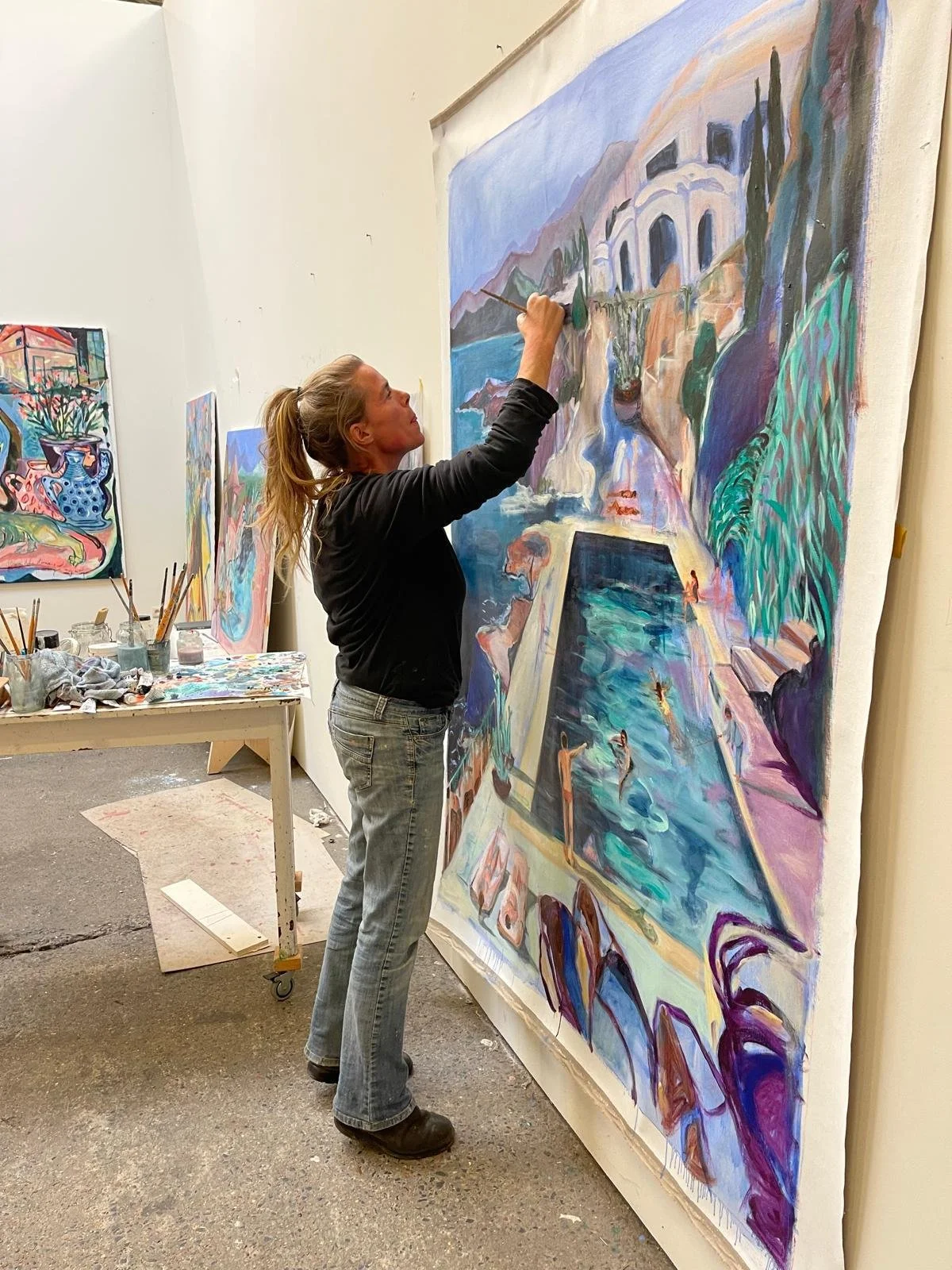 A woman painting a large colorful landscape with a pool, mountains, and trees on a canvas in an art studio.