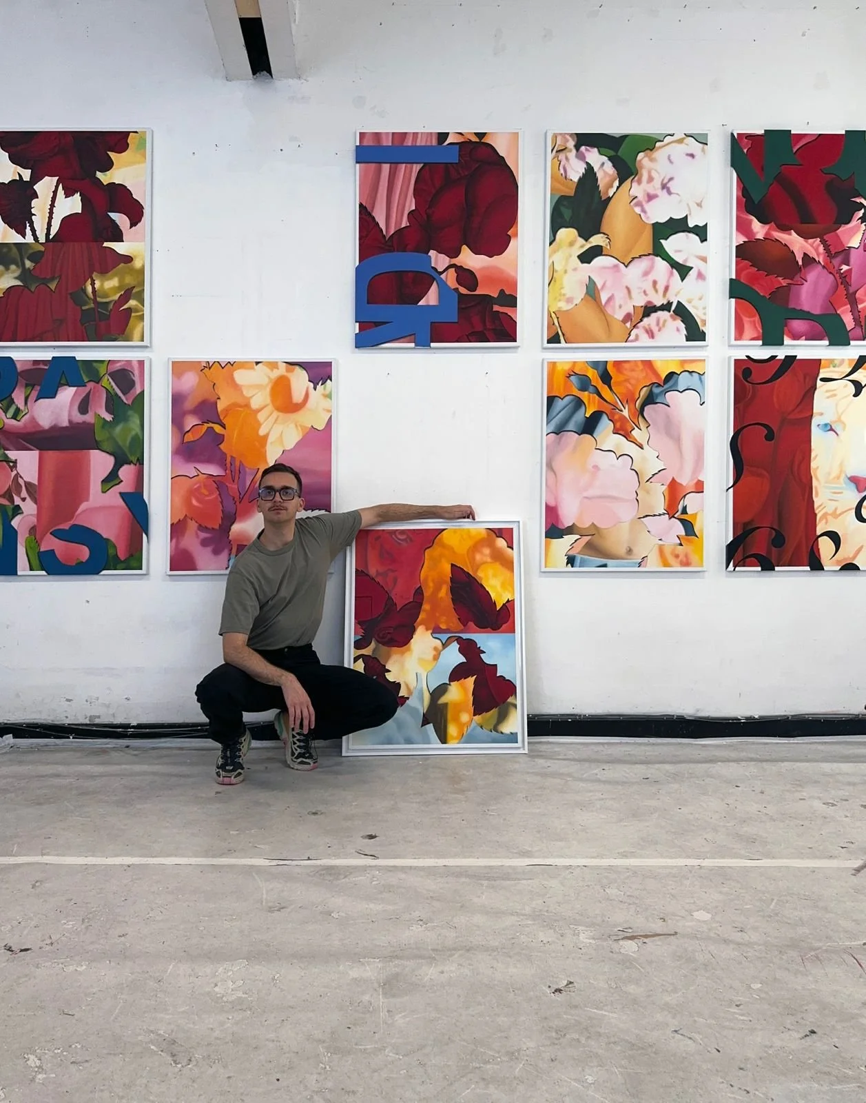 A man squatting in front of colorful abstract floral artwork on a white gallery wall.