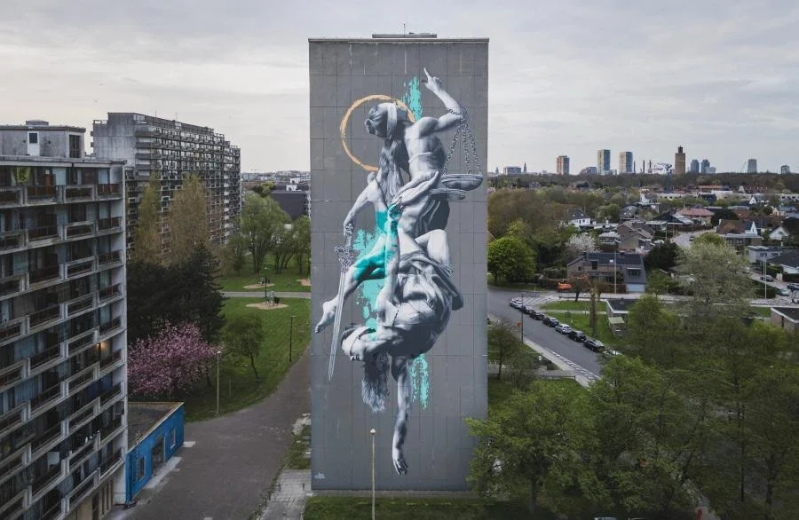 A large mural on a high building wall depicts a grayscale figure of a woman upside down, holding a sword and a scale, with abstract blue and orange splashes and a golden halo around her head. The mural is in an urban park with residential buildings, trees, and cityscape in the background.