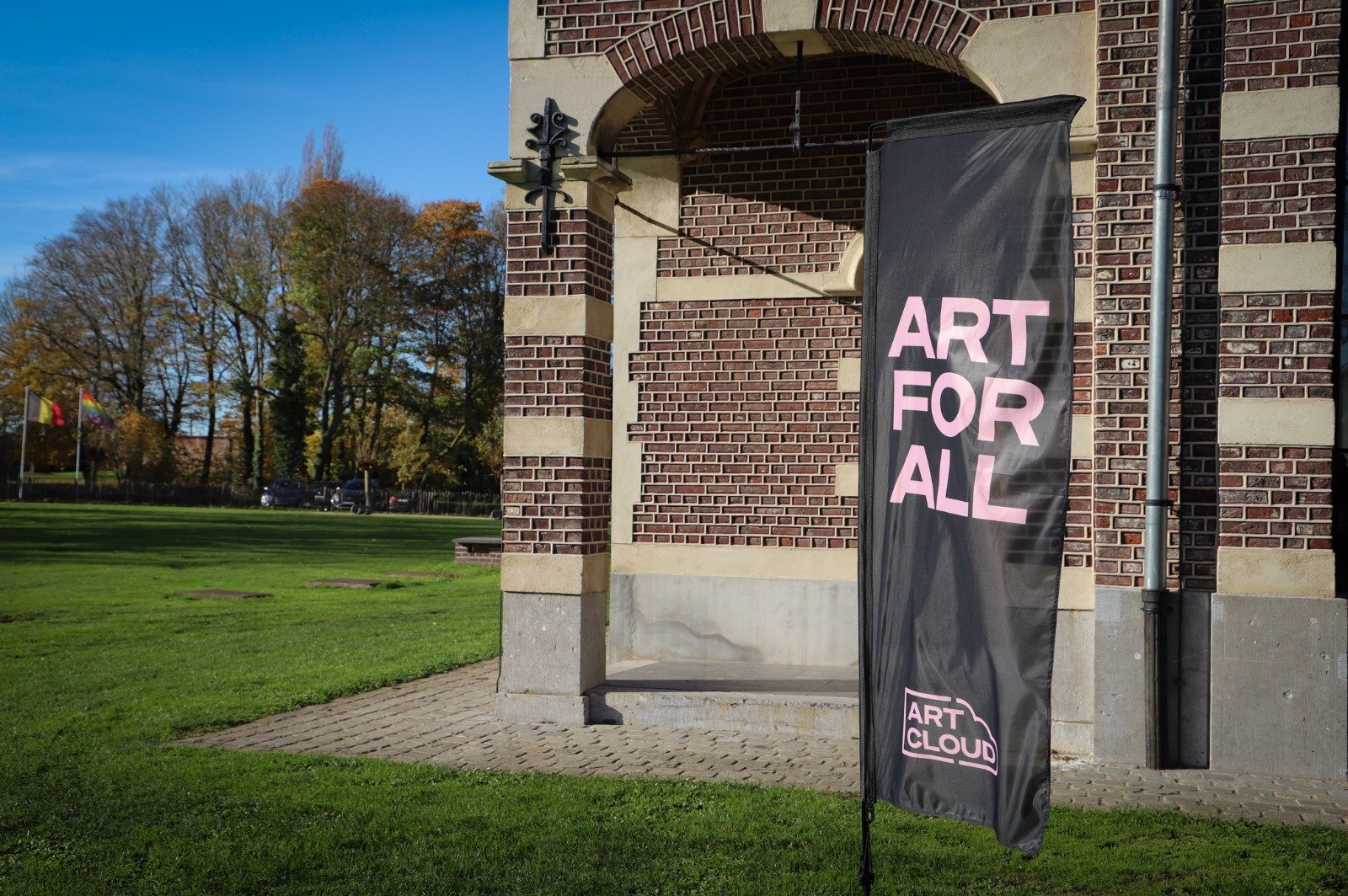 A vertical black banner with pink text reading 'ART FOR ALL' is displayed outside a brick building with an arched entrance. The banner has a logo at the bottom reading 'ART CLOUD'. The setting is a grassy area with trees in the background under a clear blue sky.