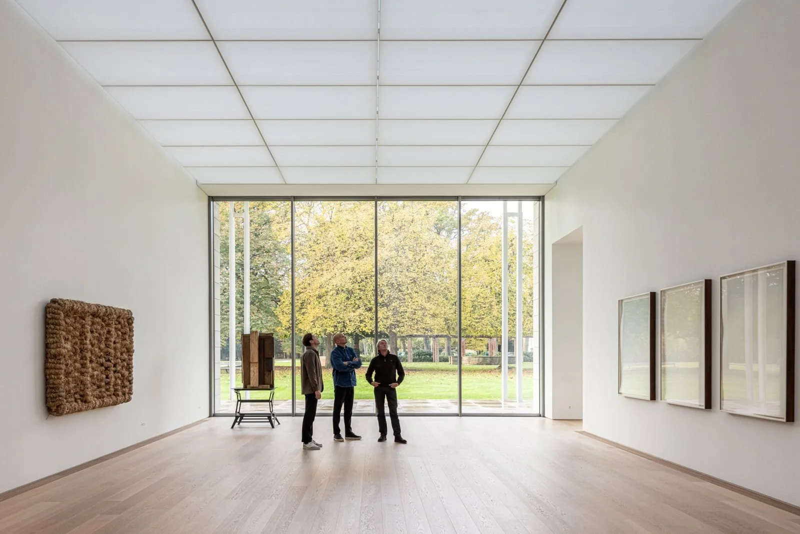 Drie mensen kijken naar kunstwerken in een witte kunstgalerij met grote ramen die uitkijken op een park met groene bomen.