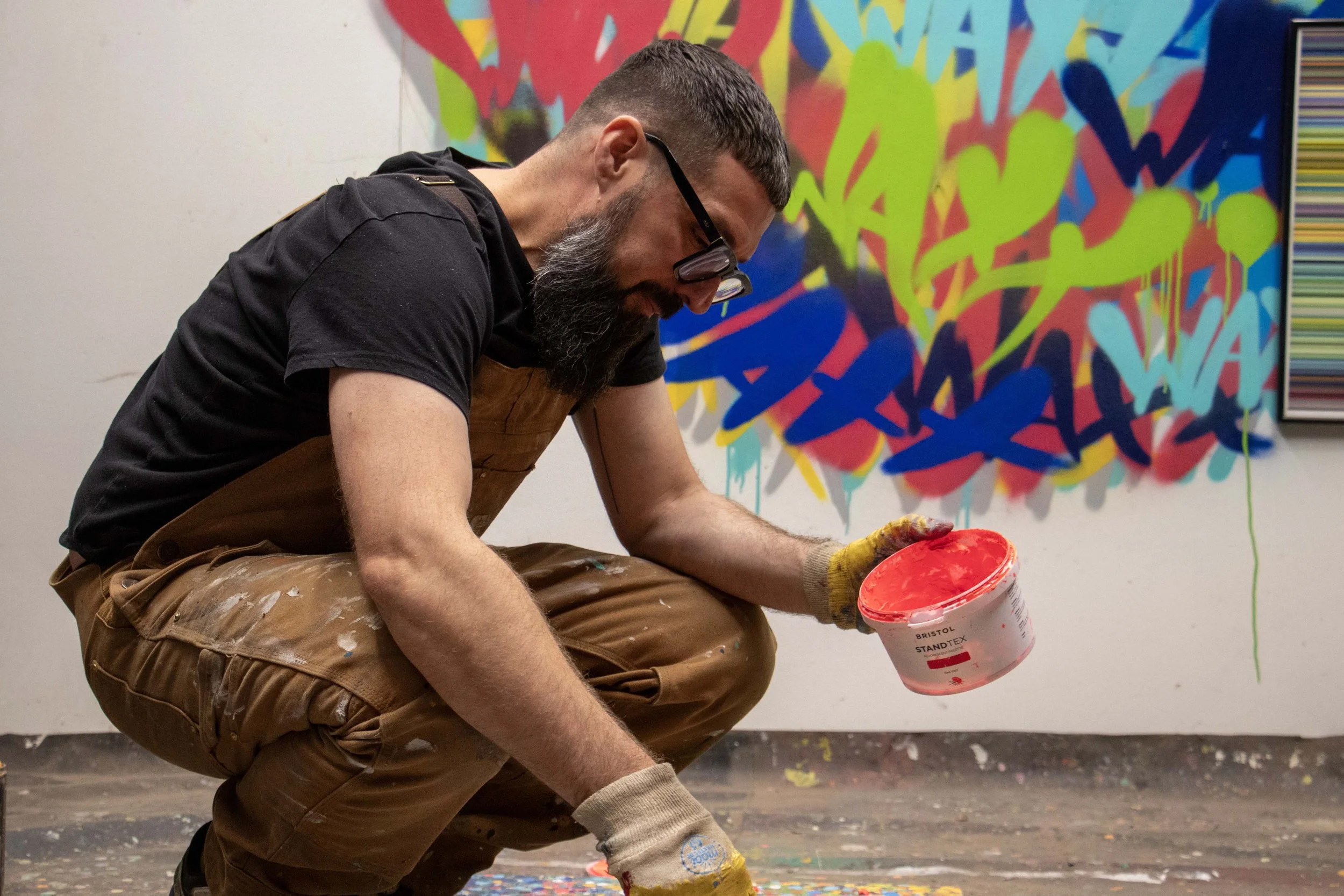 A man with glasses and a beard painting graffiti on a wall. He is kneeling, wearing a brown apron and gloves, holding a bucket of red paint. The graffiti in the background features bright, colorful abstract shapes.