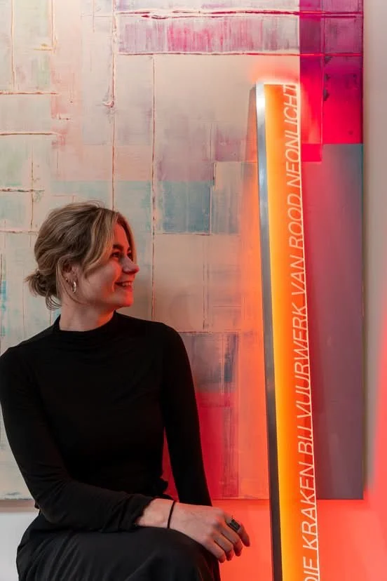 A woman with short blonde hair wearing a black long-sleeve top sits in front of a colorful abstract wall and a vertical neon sign with red-orange glow.
