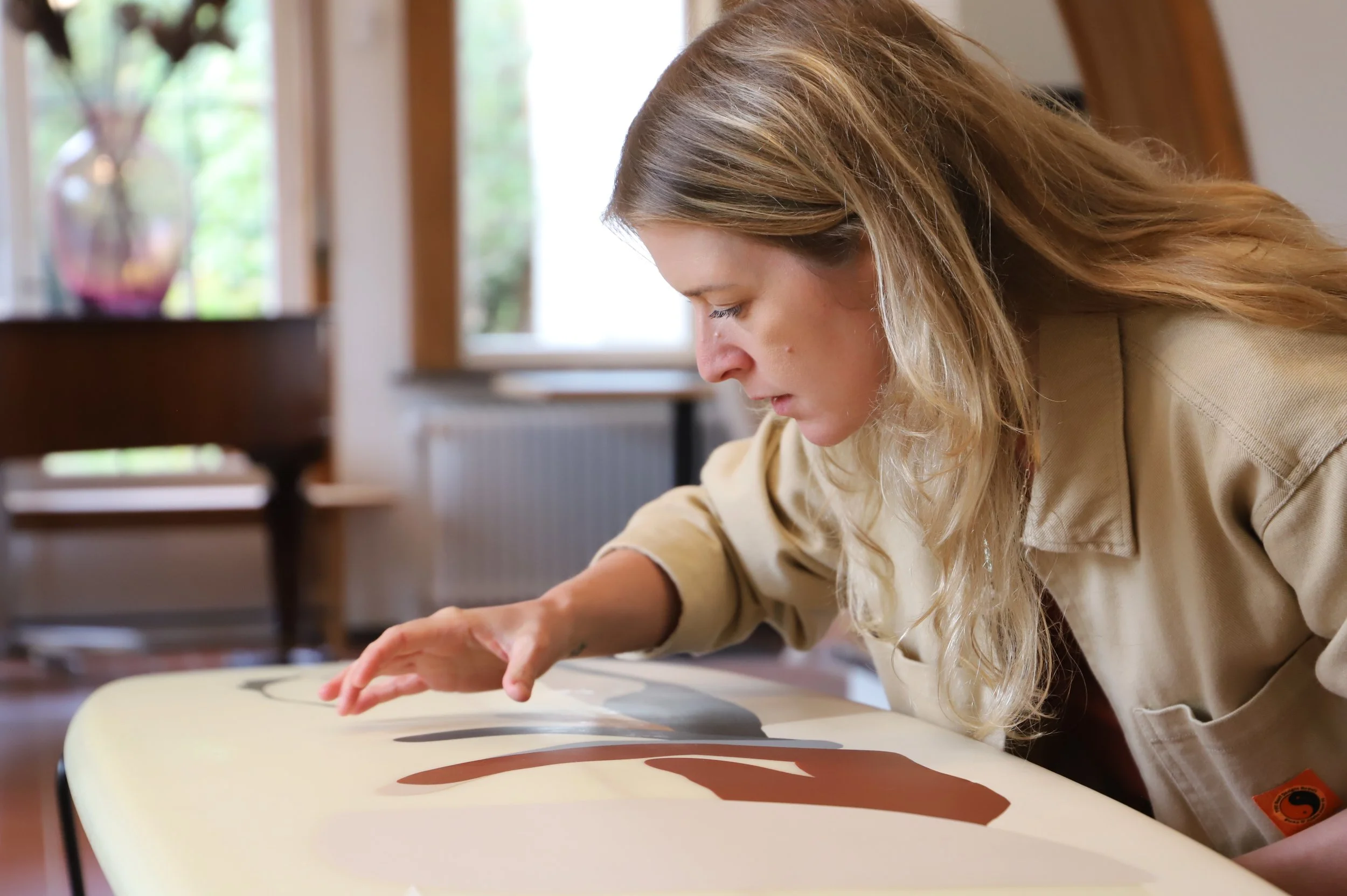 Young woman wearing a beige jacket drawing on a surfboard at a table in a bright room with large windows and potted plants.