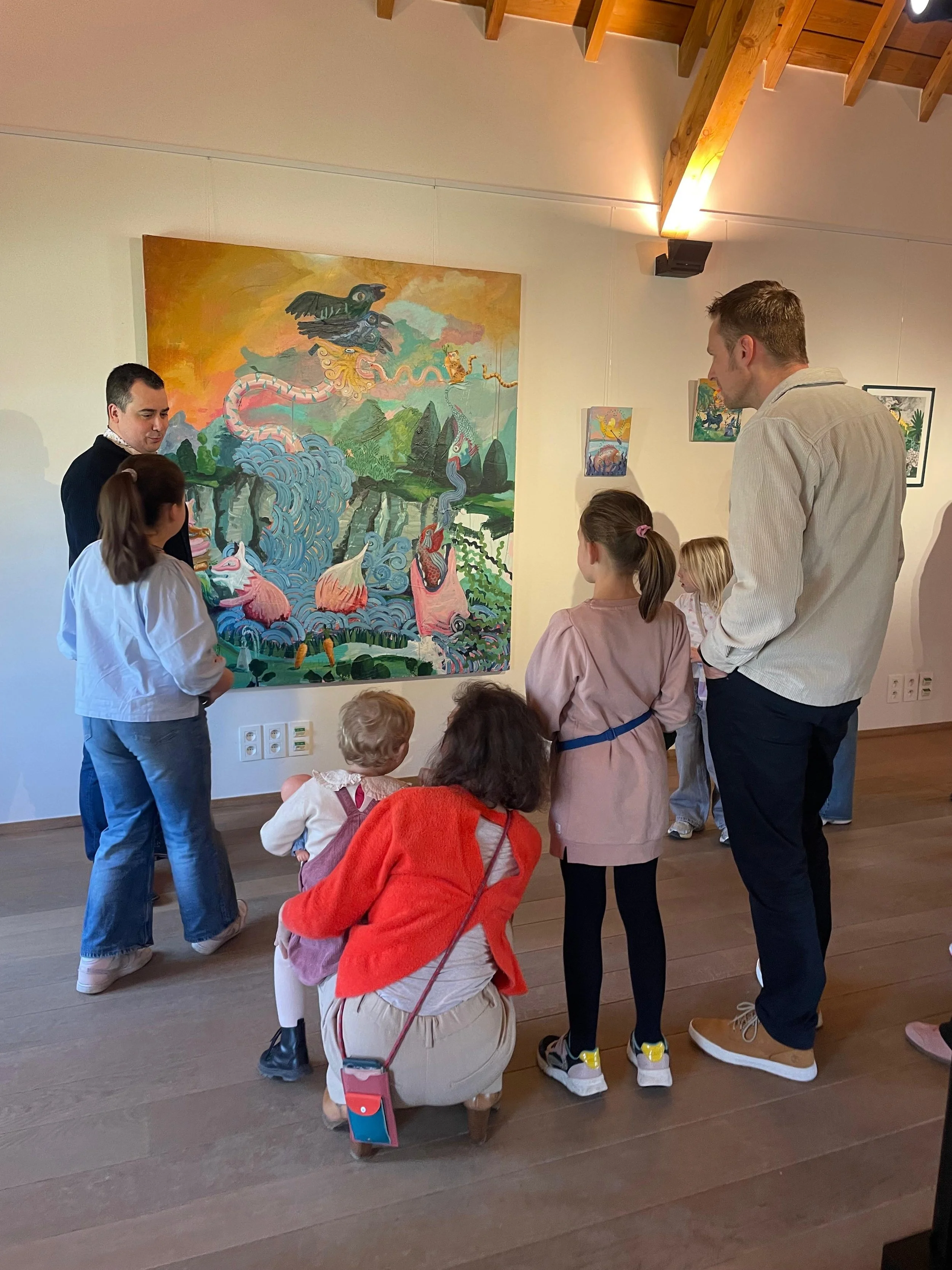 People, including children and adults, gathered in an art gallery watching a colorful painting of mythical creatures and landscape, with wooden ceiling beams overhead.