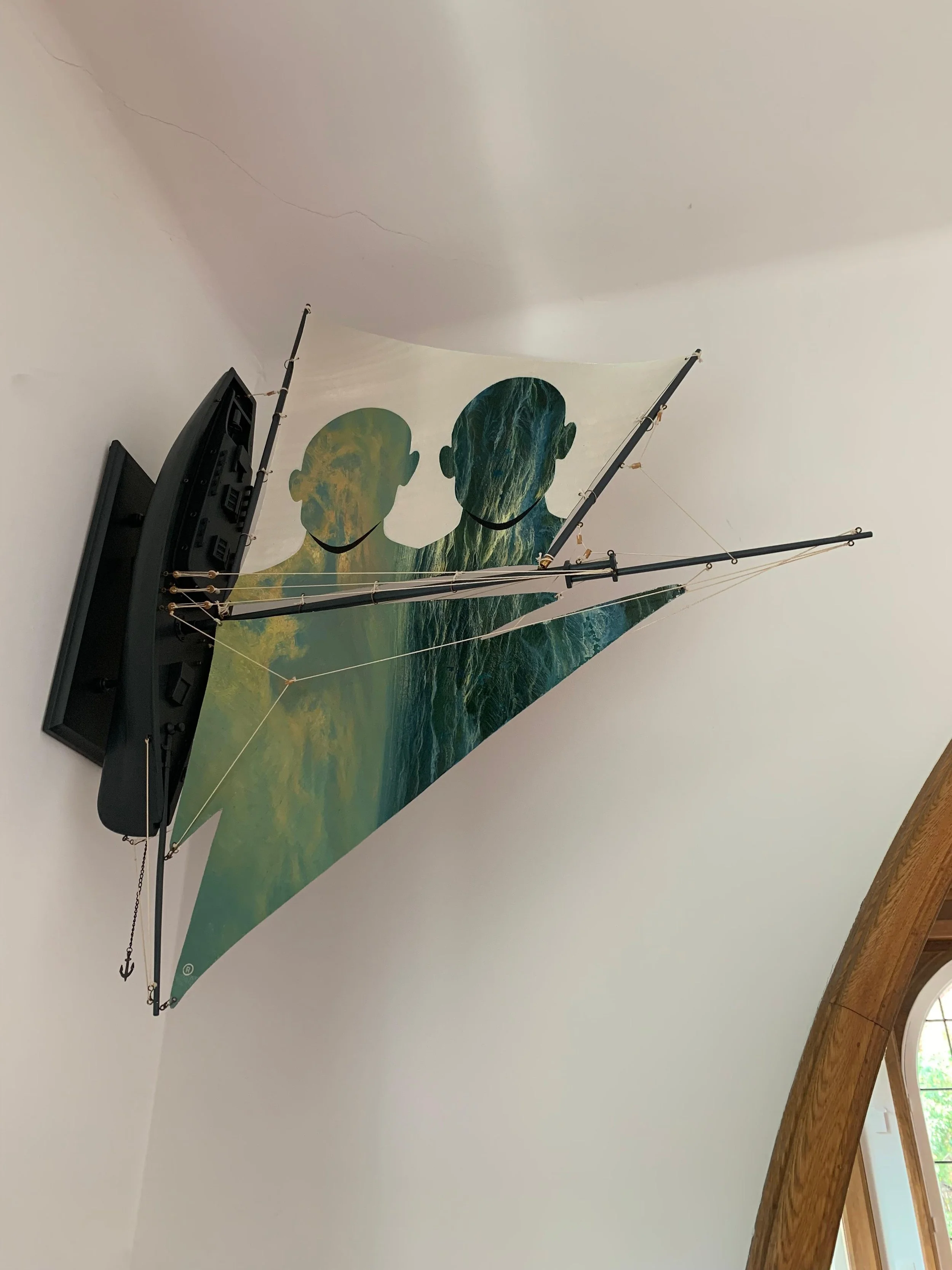 A model sailboat with a colorful sail featuring an artistic design of two faceless figures, mounted on a white wall near a wooden window frame.