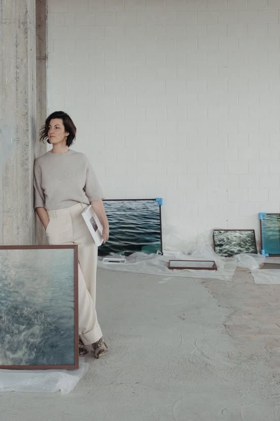 A woman standing in an art studio or gallery, surrounded by framed photographs or artwork of water scenes leaning against the wall and on the floor.