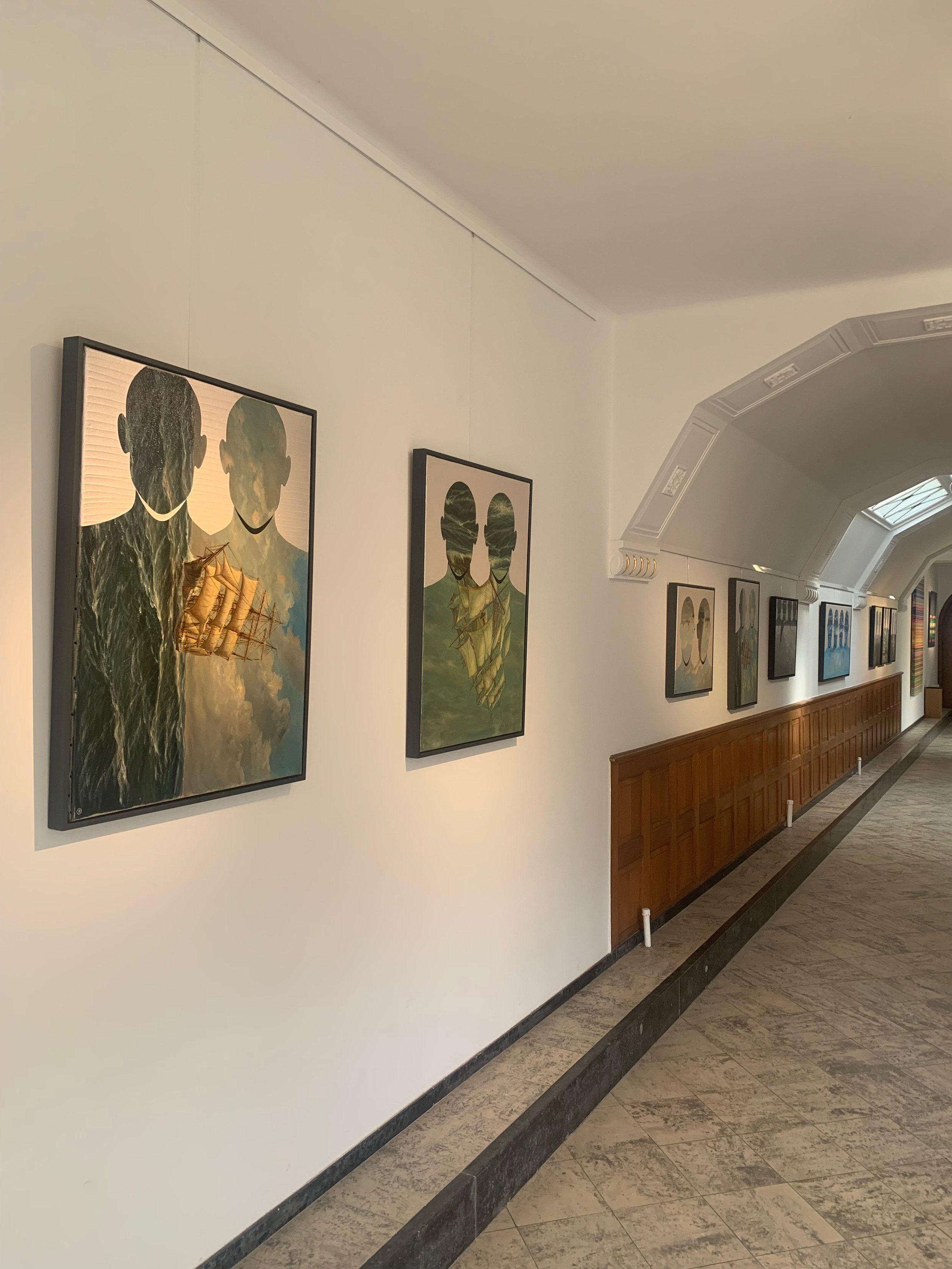 Gallery hallway with framed artwork featuring silhouettes of two people over water scenes, wooden wainscot and arched ceiling.
