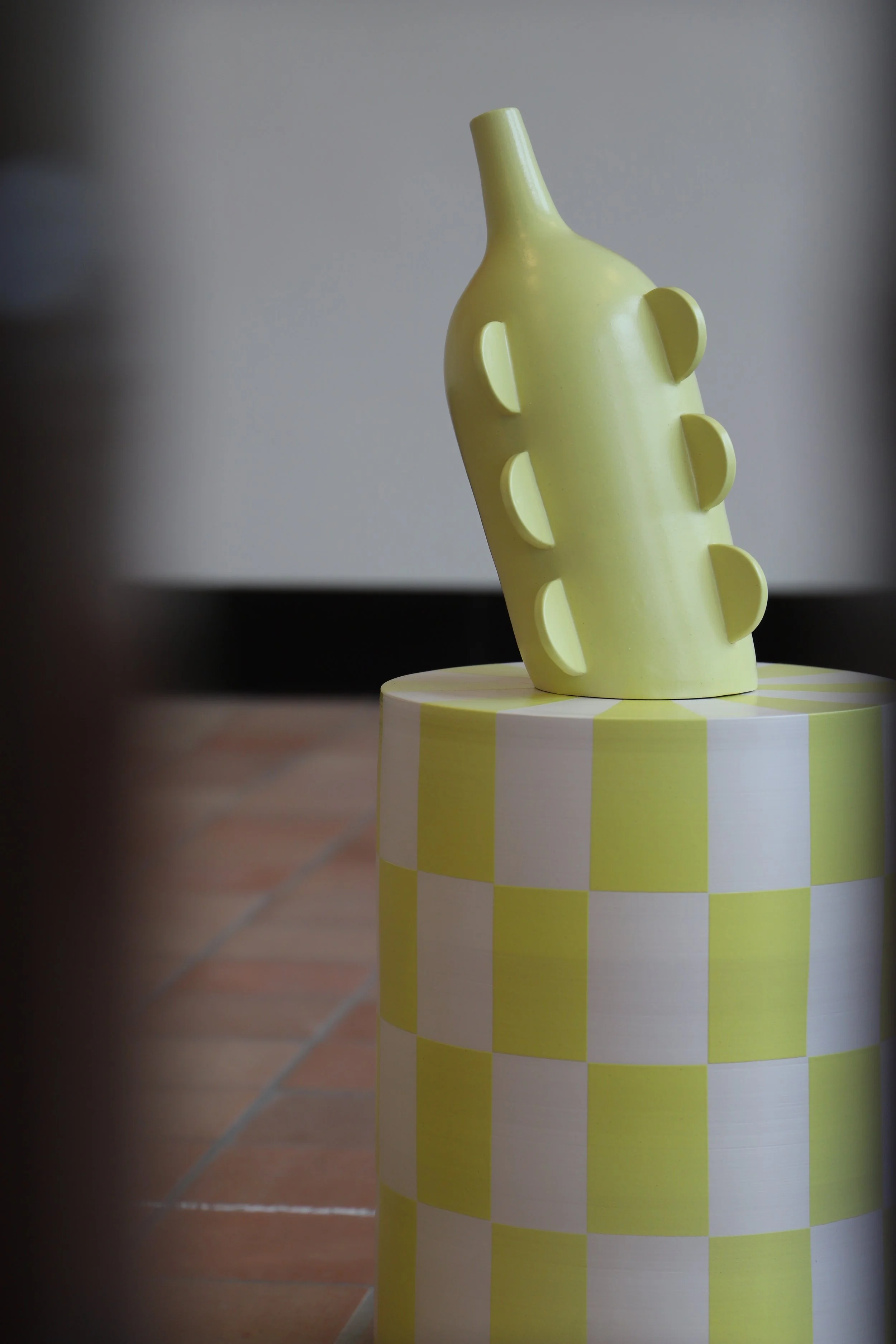A yellow decorative vase with abstract design elements sits on a yellow and white checkered pedestal.