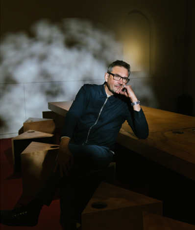 A man with glasses and a beard sitting at a wooden table in a dimly lit room with a cloud-like smoke or mist behind him, resting his chin on his hand and smiling.