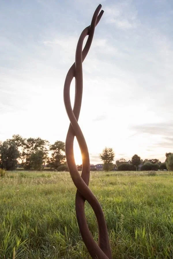 Tall outdoor sculpture made of intertwined, curving metal bars in a grassy field with trees and a sunset in the background.