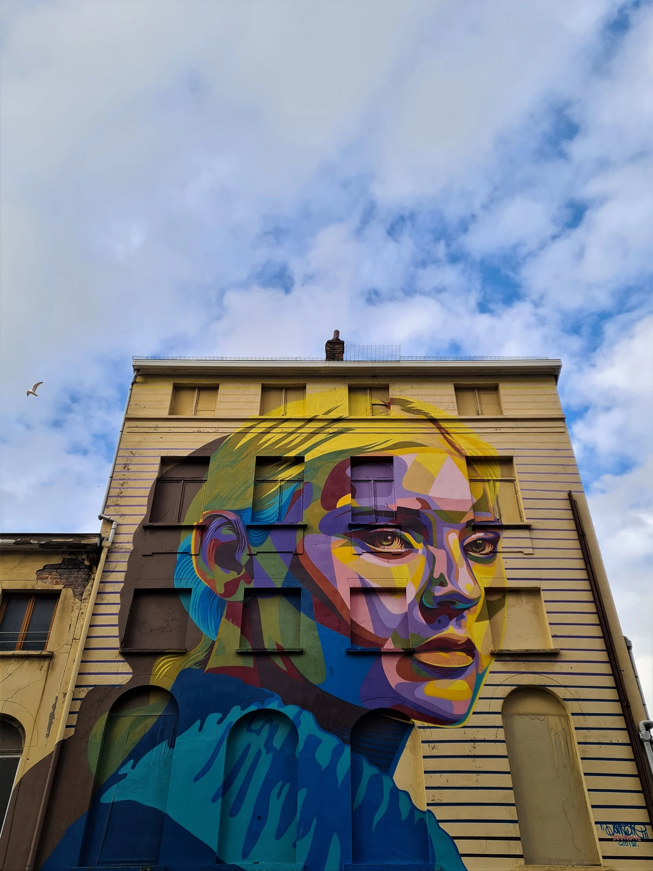 Colorful mural of a woman's face on the side of a multi-story building against a blue sky with clouds.