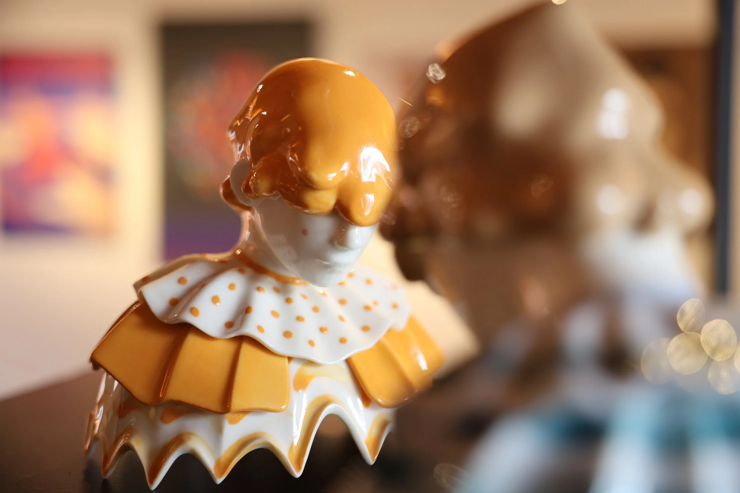 Twee argitektonische beeldjes van clowns of circusfiguren, gemaakt van glanzend keramiek, met de voorste in witte en gele details, en de achtergrond iets wazig.