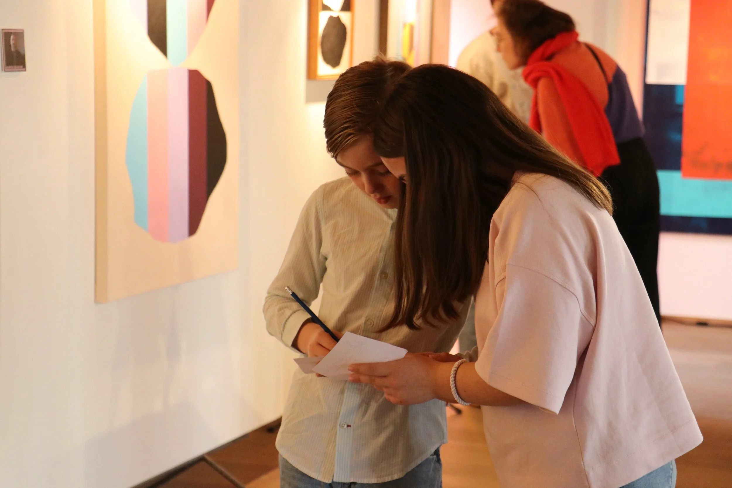 Twee jonge vrouwen kijken samen op een stuk papier in een kunsttentoonstelling, terwijl andere mensen kunst bekijken op de achtergrond.