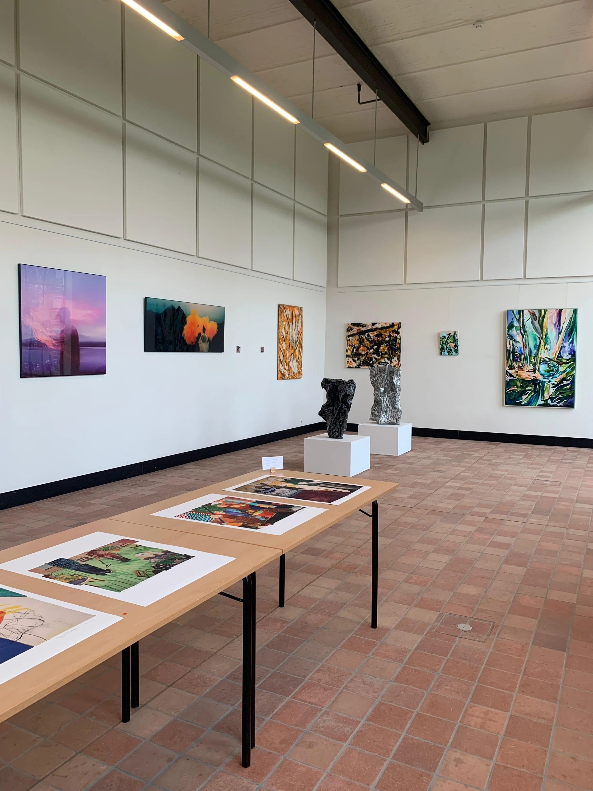 Art gallery with colorful paintings on the wall, artist sculptures on pedestals, and artwork displayed on tables and walls in a spacious room with brick floors and modern lighting.