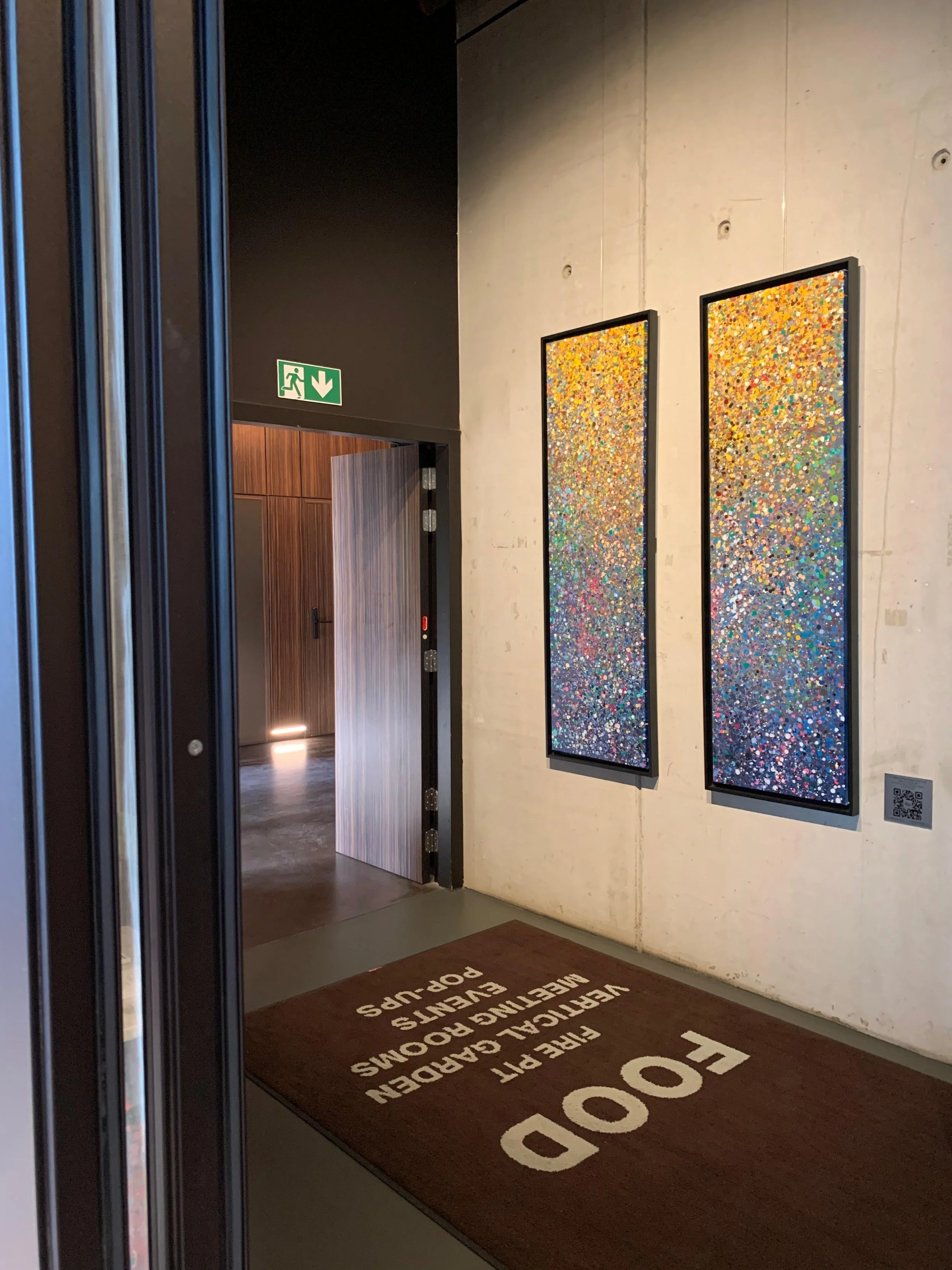 Interior of a building with a door slightly open, leading to a hallway. Two colorful abstract paintings hang on a plain wall. Emergency exit sign above the doorway. Doormat on the floor reads "FODO" and additional safety instructions.