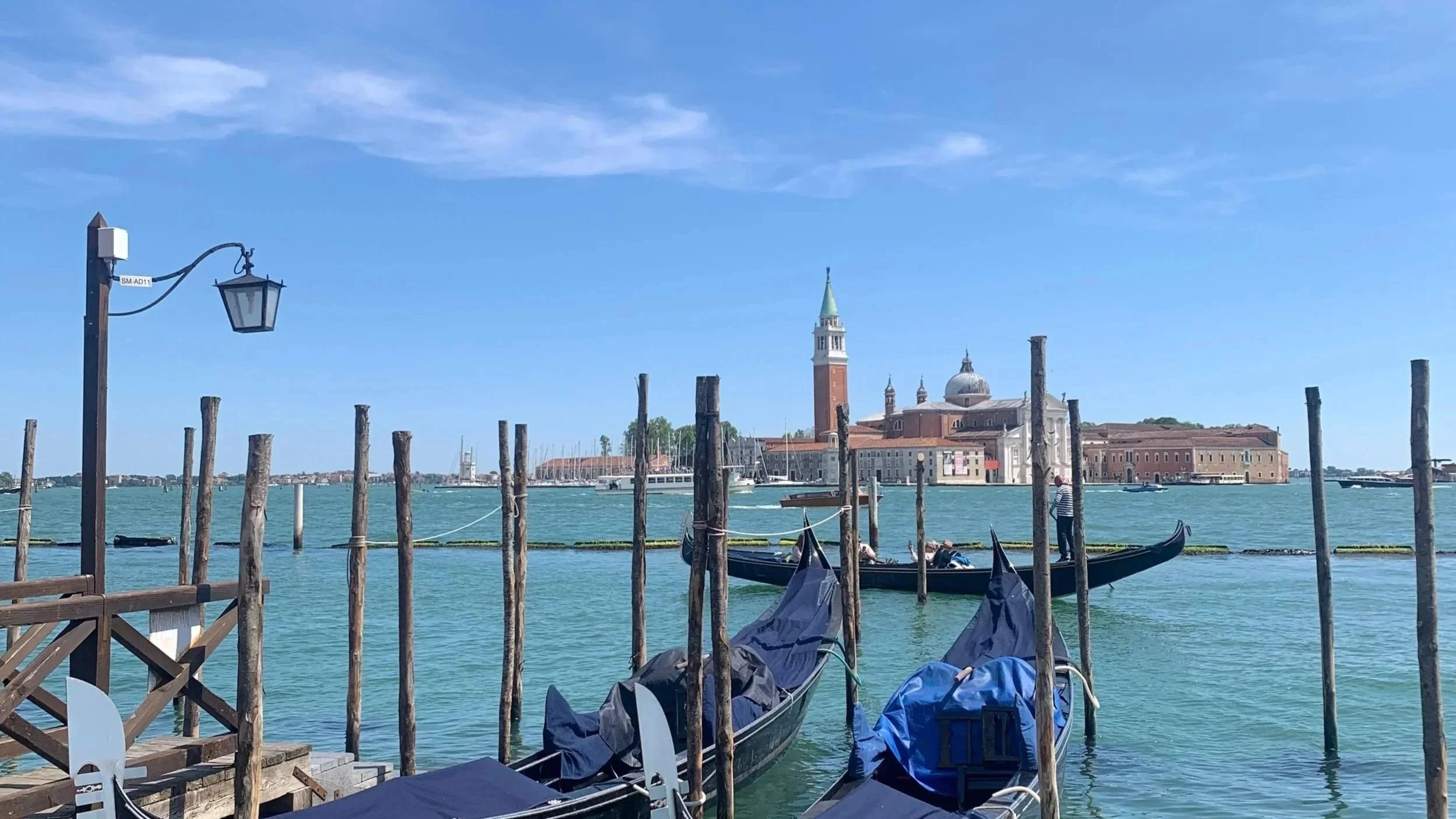 View of a canal in Venice with gondolas docked, wooden posts in water, and historic buildings including the San Giorgio Maggiore church in the background under a blue sky.