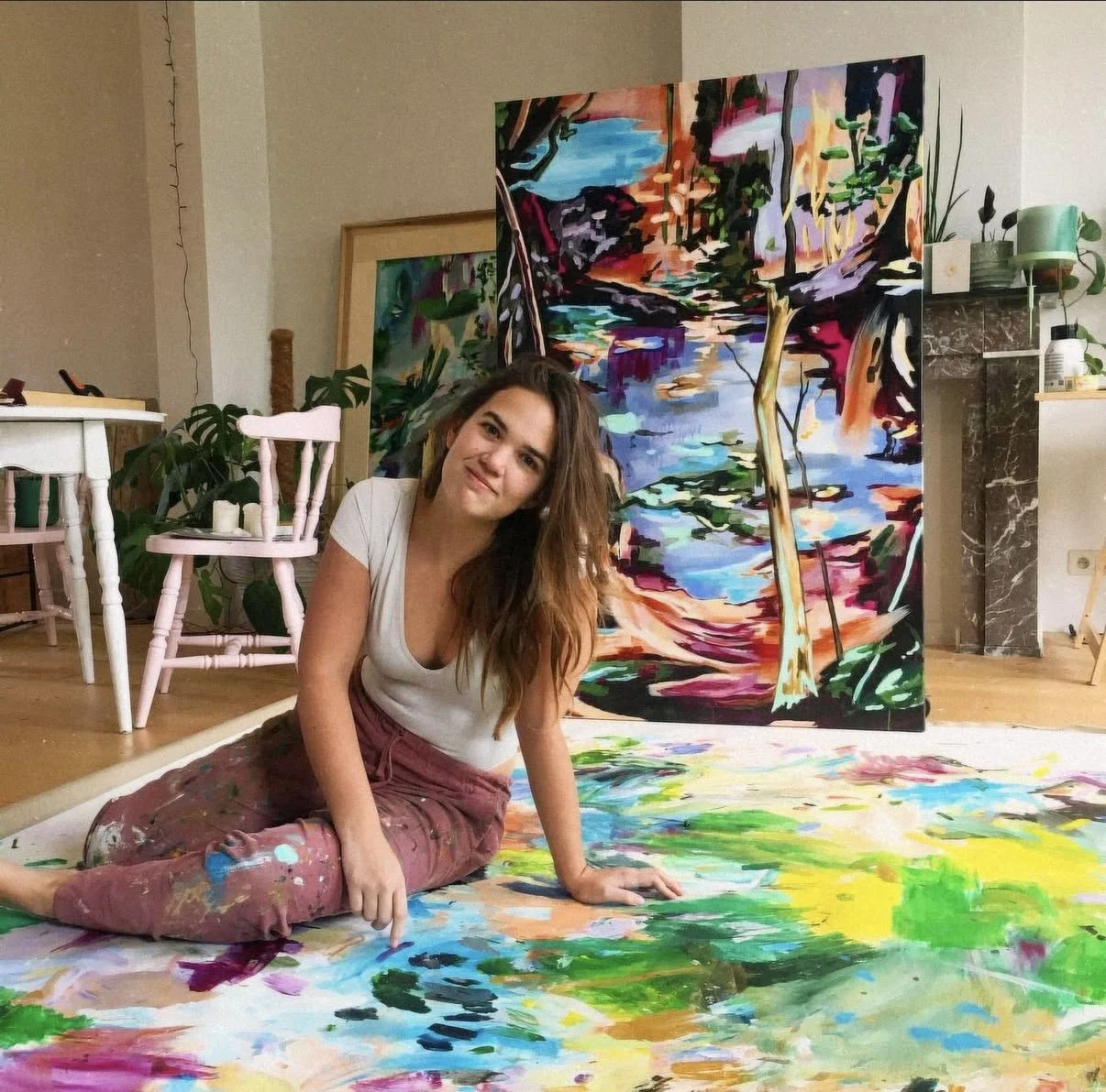 Young woman with long hair smiling, sitting on a large canvas with colorful abstract paint, in front of a vibrant abstract landscape painting in a creative indoor space.