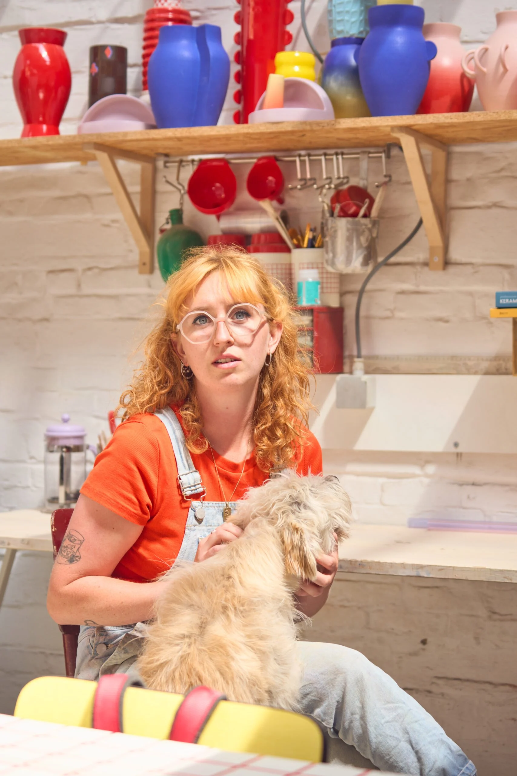 A red-haired woman wearing glasses, a red shirt, and denim overalls sitting at a table holding a fluffy light-colored dog, with a kitchen or workshop background featuring shelves with colorful vases and various tools.
