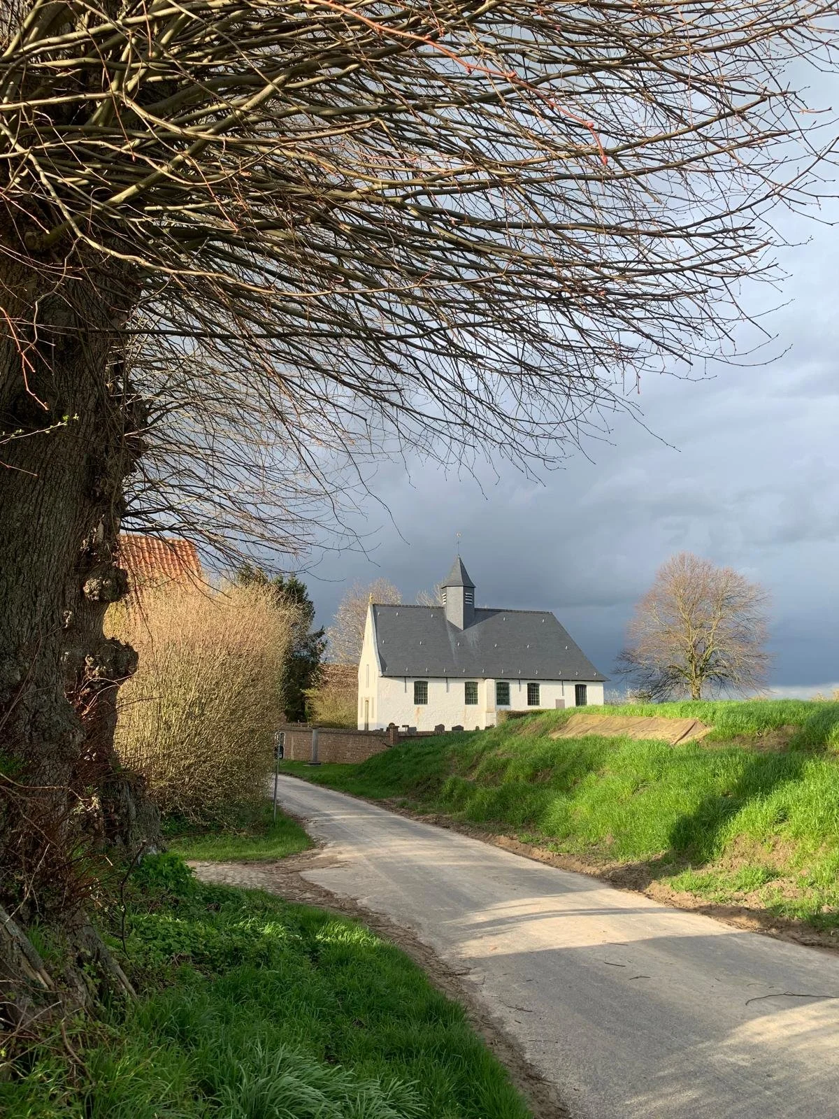 A country road with green grass on both sides, a white church in the distance with a dark roof and steeple, surrounded by leafless trees, under a cloudy sky.