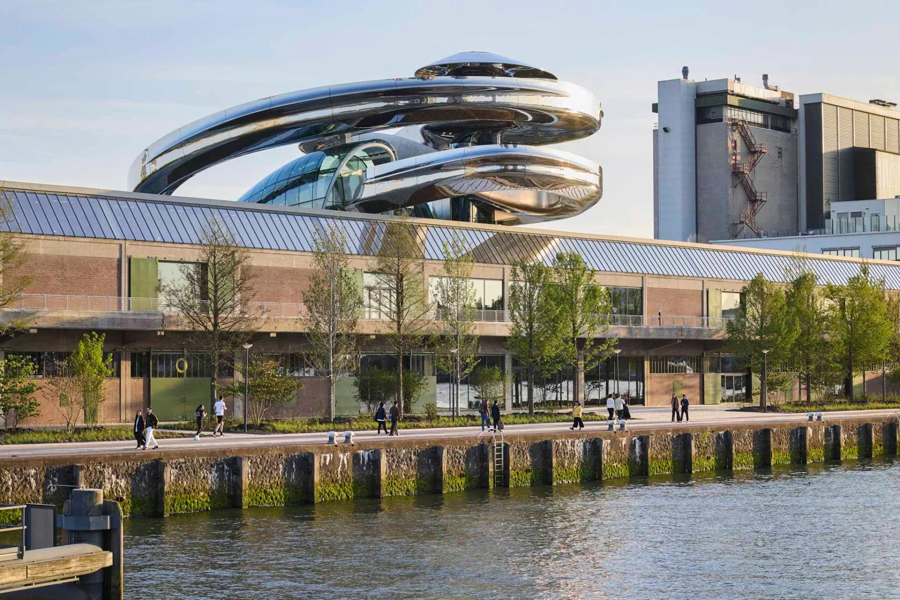 Modern gebouw met een glanzende, ronde, snoer-achtige structuur op het dak, gelegen aan een rivier, met mensen die langs de promenade wandelen.