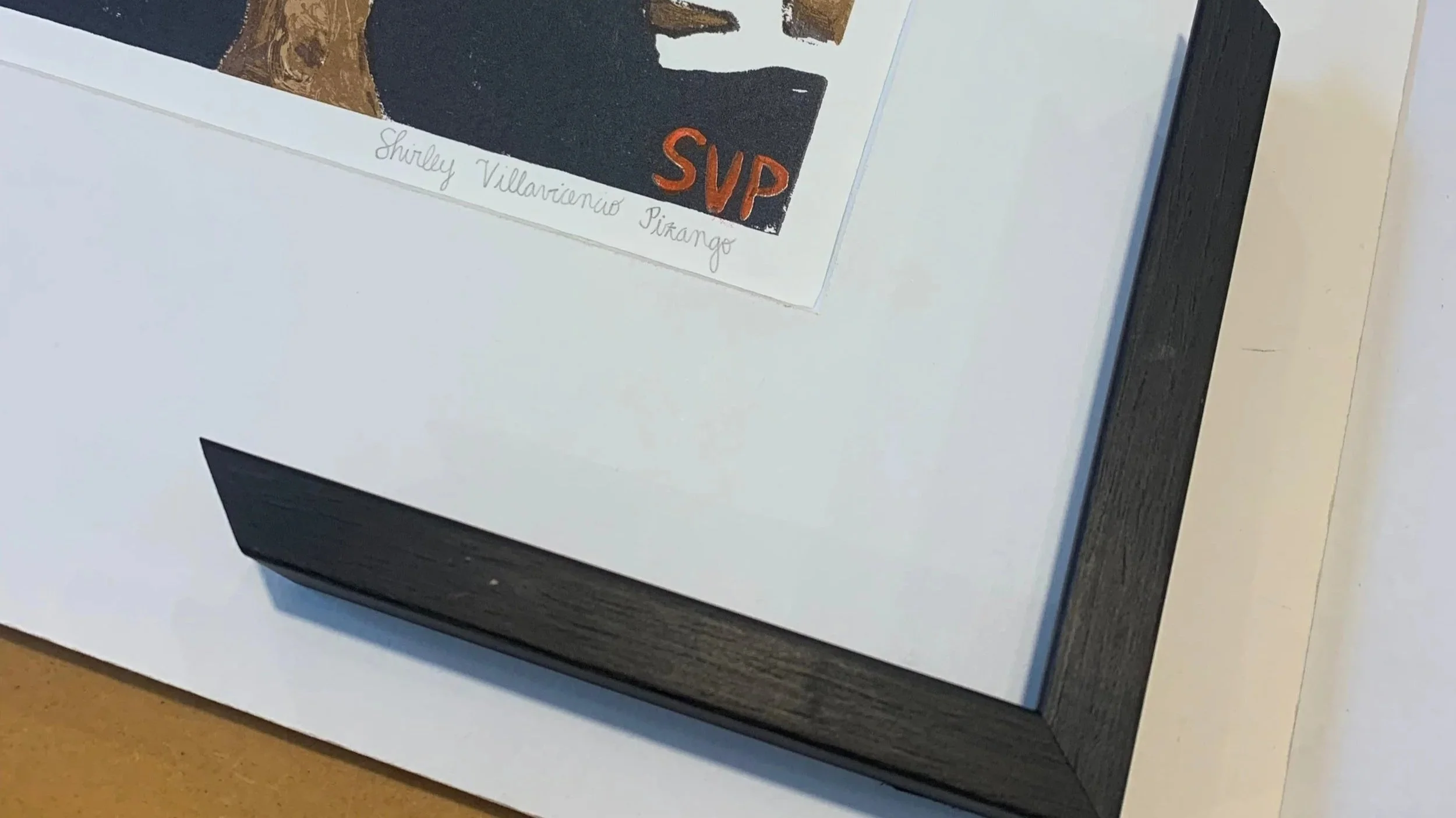 Close-up of a framed artwork with the artist's signature 'Shirley Villavicencio Pizango' in fine script, and a section of the artwork showing black, brown, and orange colors with the initials 'SVP' in orange.